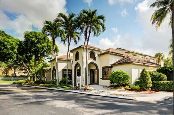 A large, multi-story house with white walls, brown roof, and arched windows, surrounded by palm trees and lush greenery in a sunny, suburban neighborhood.