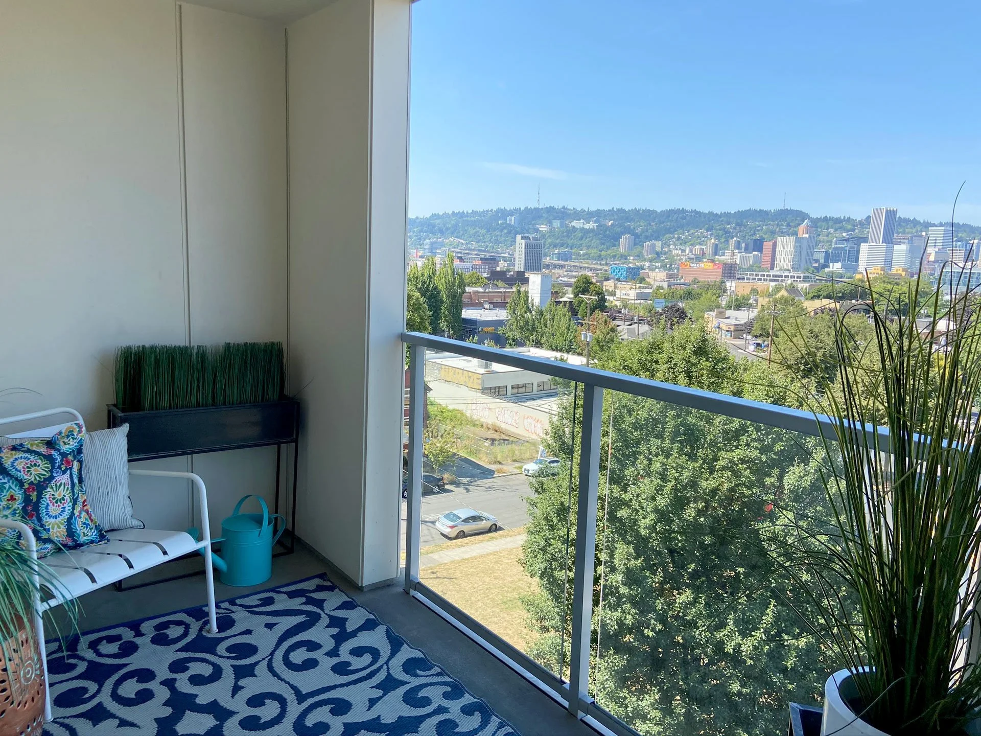 A balcony with a view of a city skyline, trees, cars, and hills in the background. The balcony has a white metal bench with colorful cushions, a blue watering can, a patterned outdoor rug, a tall potted plant, and a black rectangular planter with gre
