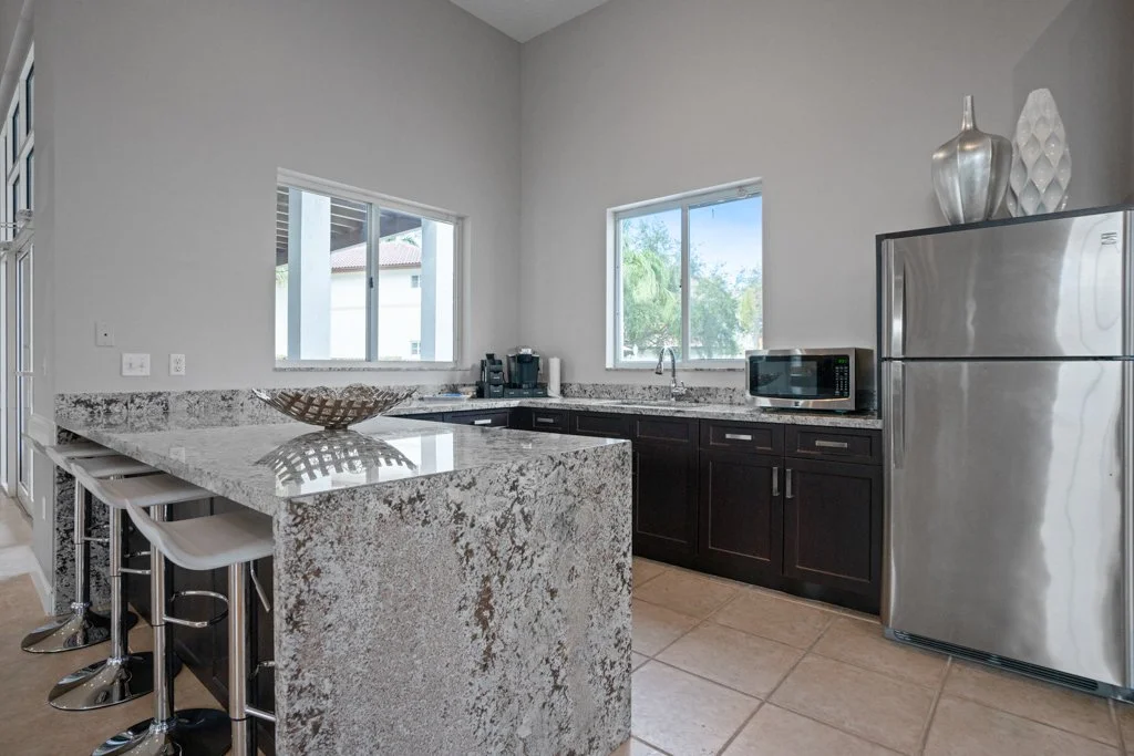 Modern kitchen with granite countertops, black cabinets, stainless steel refrigerator, microwave, and bar stools, with two windows overlooking greenery.