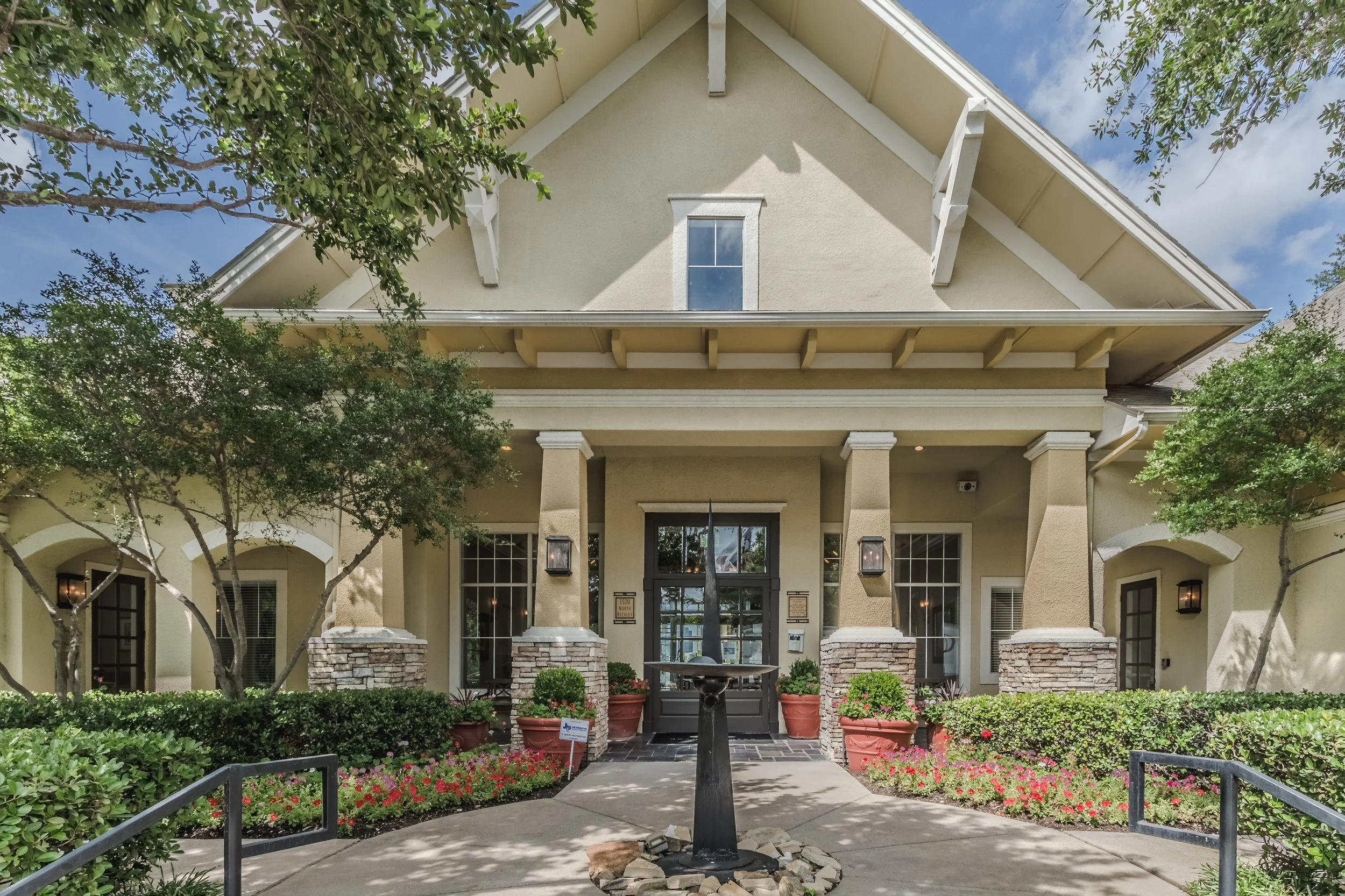 Front view of a beige house with a gable roof, decorated with white trim and columns. There is a walkway leading to the front door with a water fountain in the center, surrounded by plants and flowers.