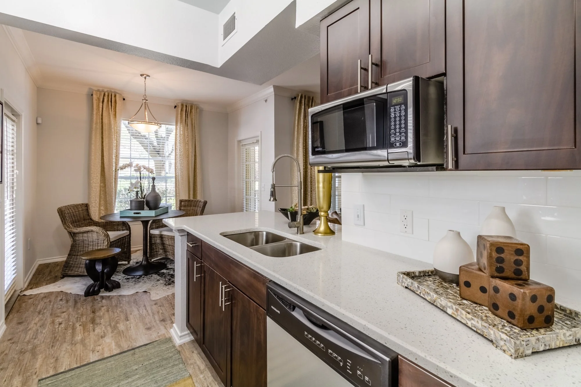Kitchen with dark wood cabinets, white countertop, stainless steel microwave, sink, and dishwasher. Decor includes vases, dice, and a gold-colored vase. Adjacent is a dining area with a round table, wicker chairs, and a window with gold curtains.