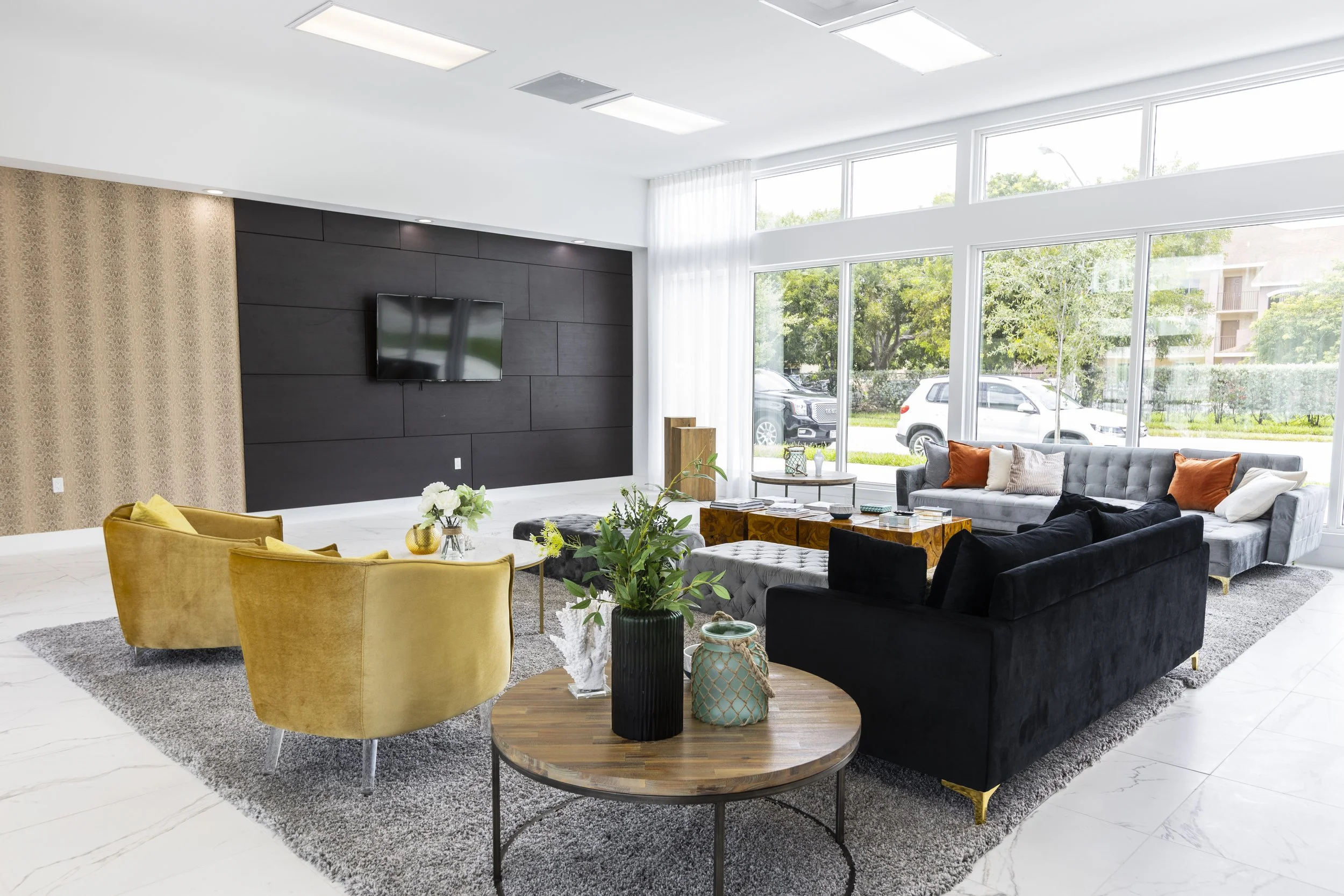 Modern living room with large windows, black, gray, and yellow sofas, a coffee table, indoor plants, and a flat-screen TV.