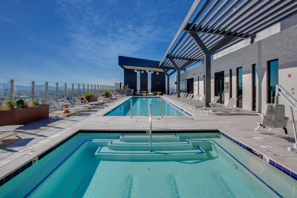 Rooftop pool area with lounge chairs, umbrellas, and a view of the city skyline under a blue sky.