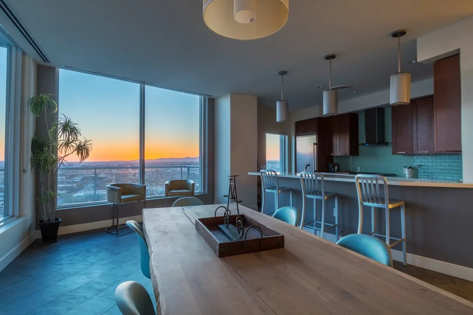 Sunset viewed from a high-rise apartment with a large window, showing the cityscape and skyline. The interior includes a dining table, chairs, bar stools at a kitchen counter, and a potted plant.