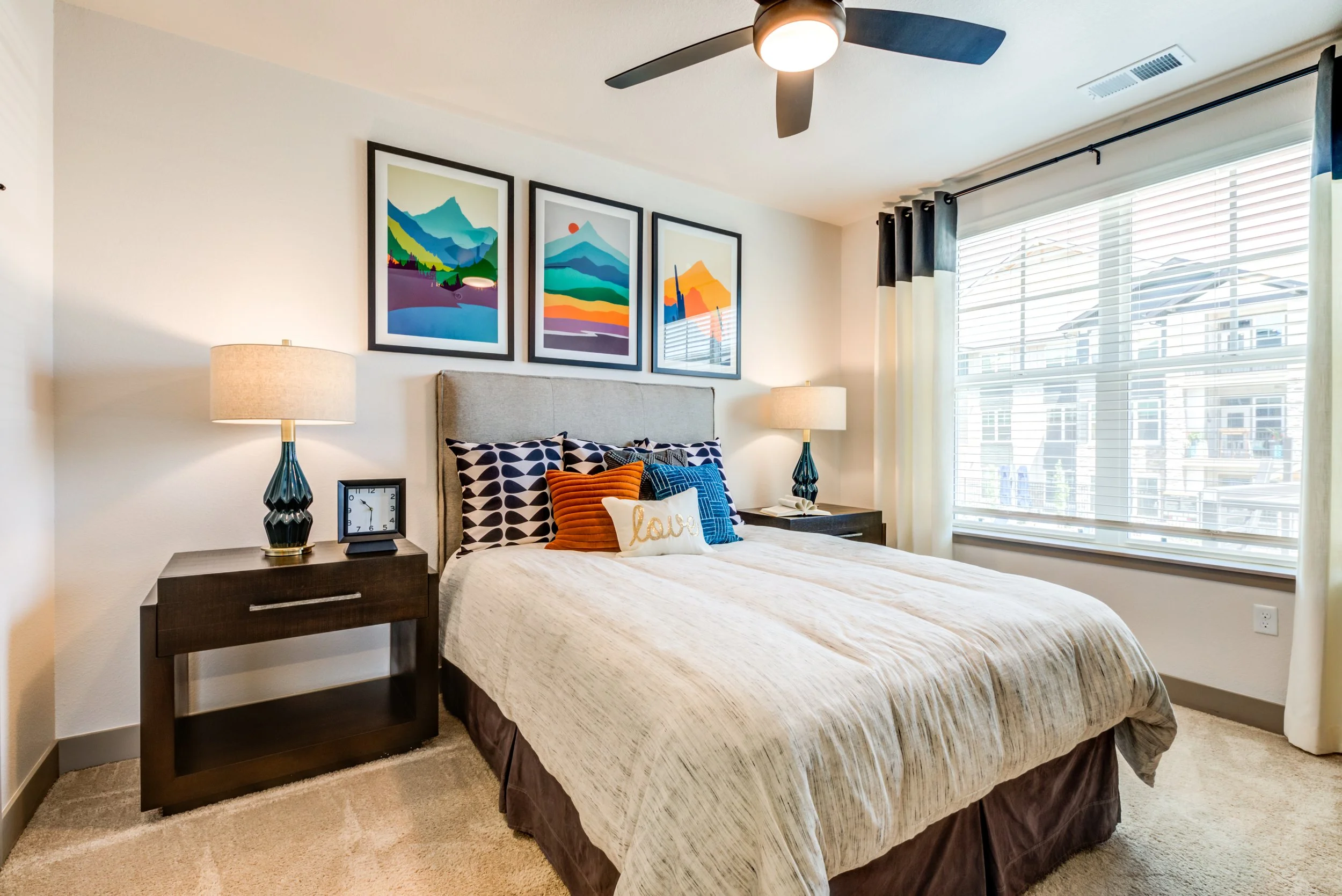 A bedroom with a bed, colorful pillows, two nightstands with lamps and clocks, artwork above the bed, a large window with blinds and curtains, and a ceiling fan.