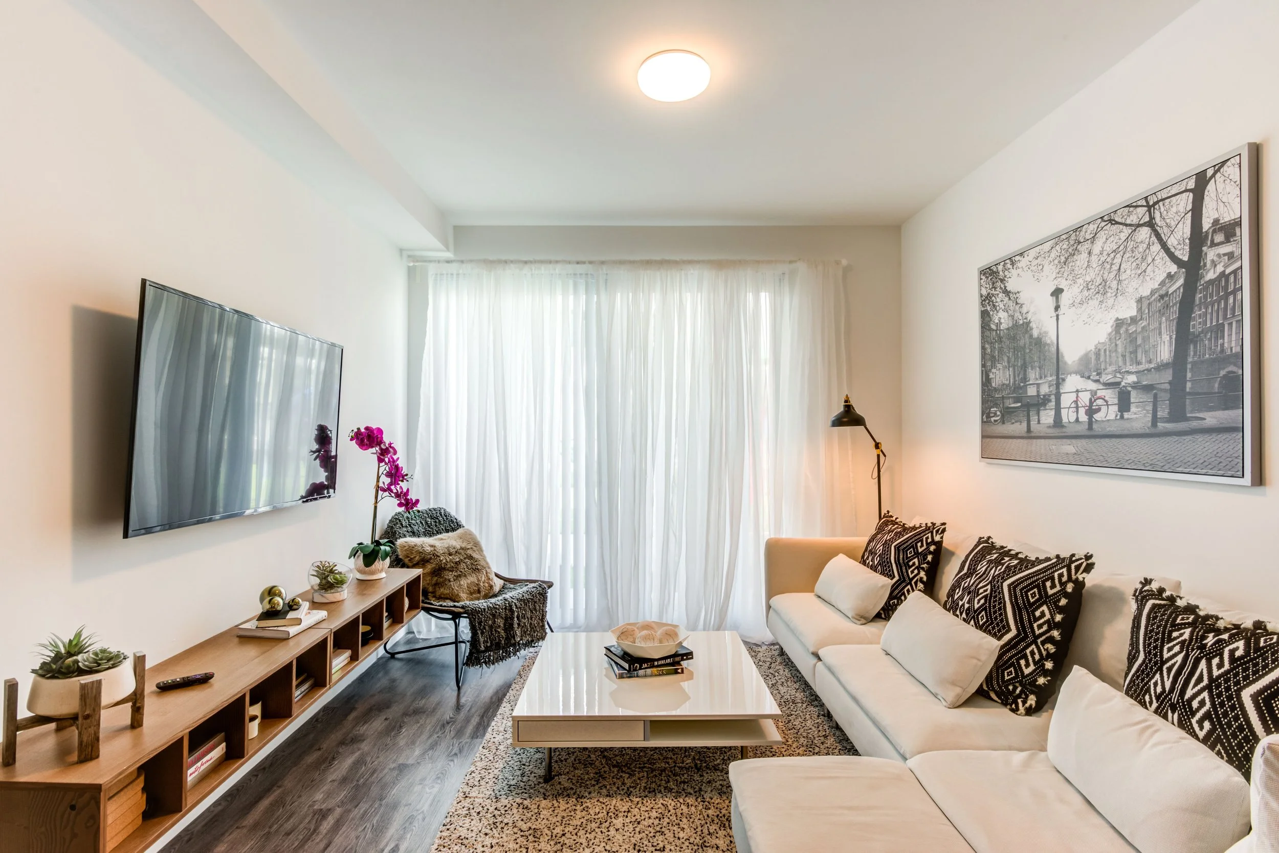 Modern living room with white sofa, patterned black and white cushions, a white coffee table, a black armchair with a throw blanket and pillows, a wall-mounted TV, houseplants, a sideboard, a floor lamp, and a black and white cityscape photograph on 