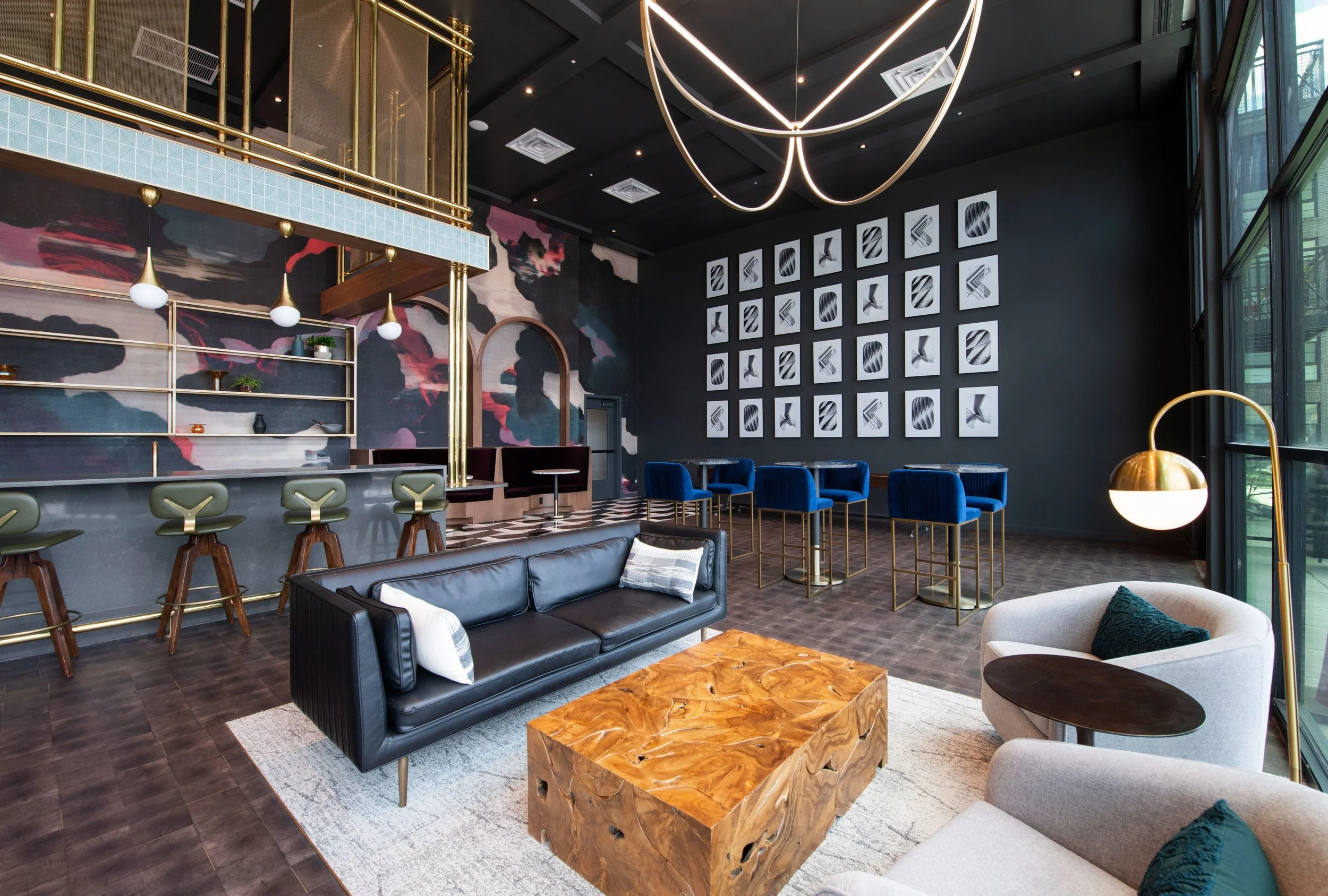 Modern lounge with black leather sofa, white armchair, wooden coffee table, blue velvet chairs, bar area with green chairs, decorative wall art, and large windows.