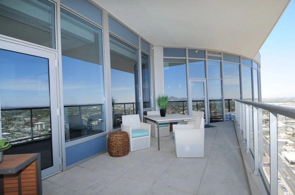 A spacious outdoor balcony with glass walls and a city view, featuring a small table with a potted plant and three chairs, some wicker and some with cushions.