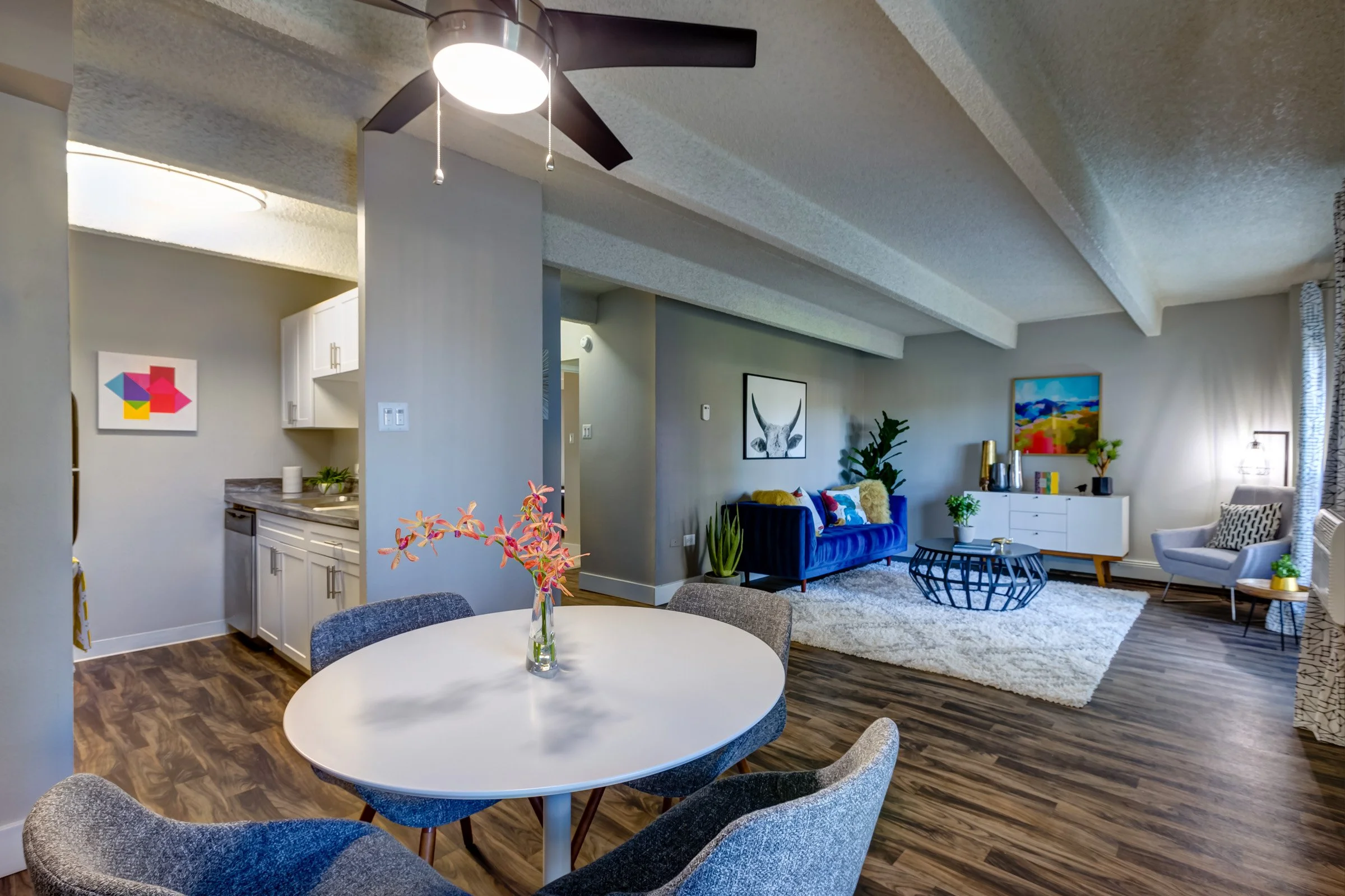 Living room and dining area with modern decor, blue velvet sofa, white rug, artwork, plants, and a round white dining table with gray chairs.