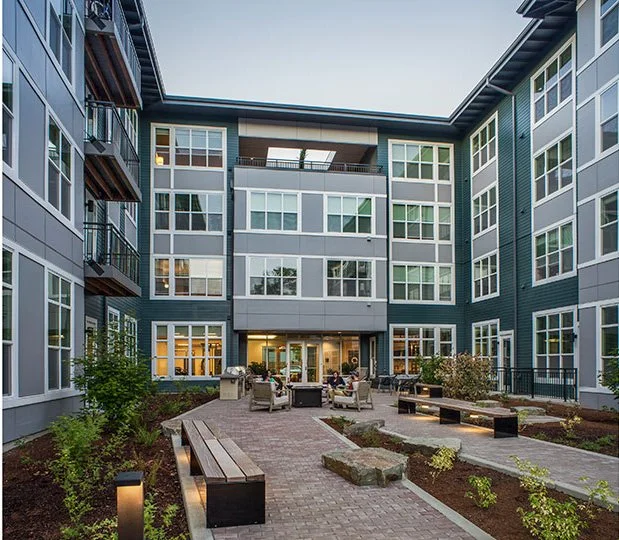 Courtyard in front of a modern apartment complex with benches, plants, and outdoor seating, during early evening.