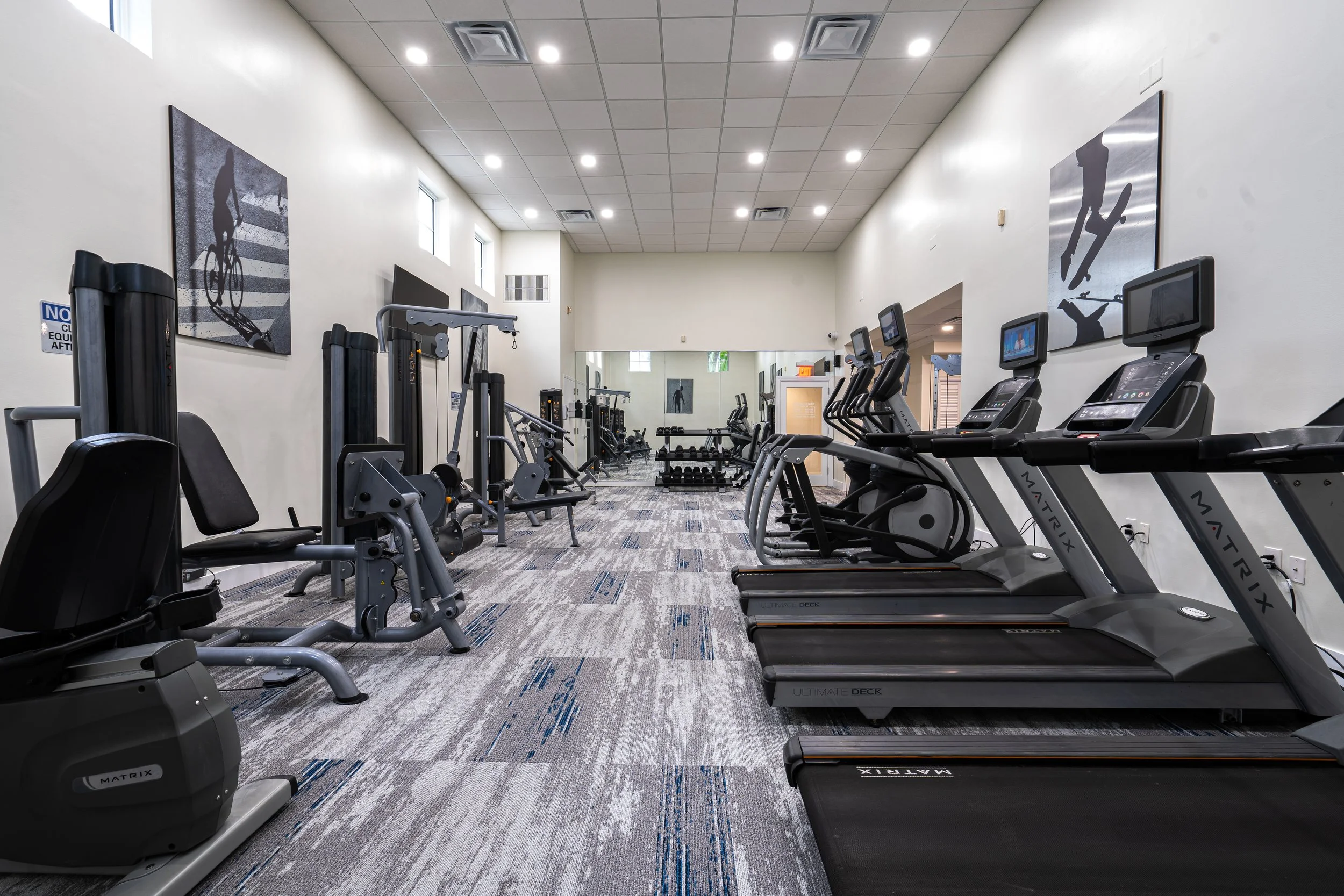 A modern gym with cardio equipment including treadmills, ellipticals, and weight machines, with artwork of athletic figures on the walls.