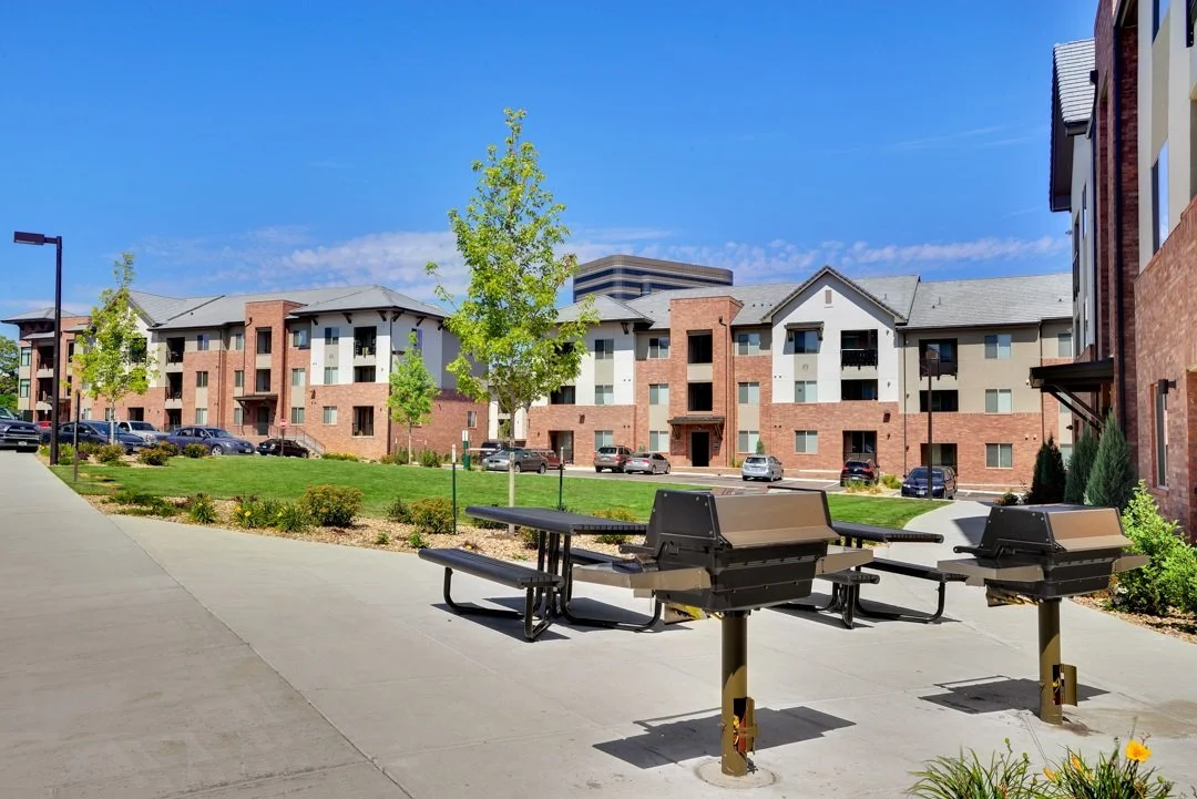 Outdoor view of a residential apartment complex with a landscaped courtyard, picnic tables, and parked cars under a blue sky.