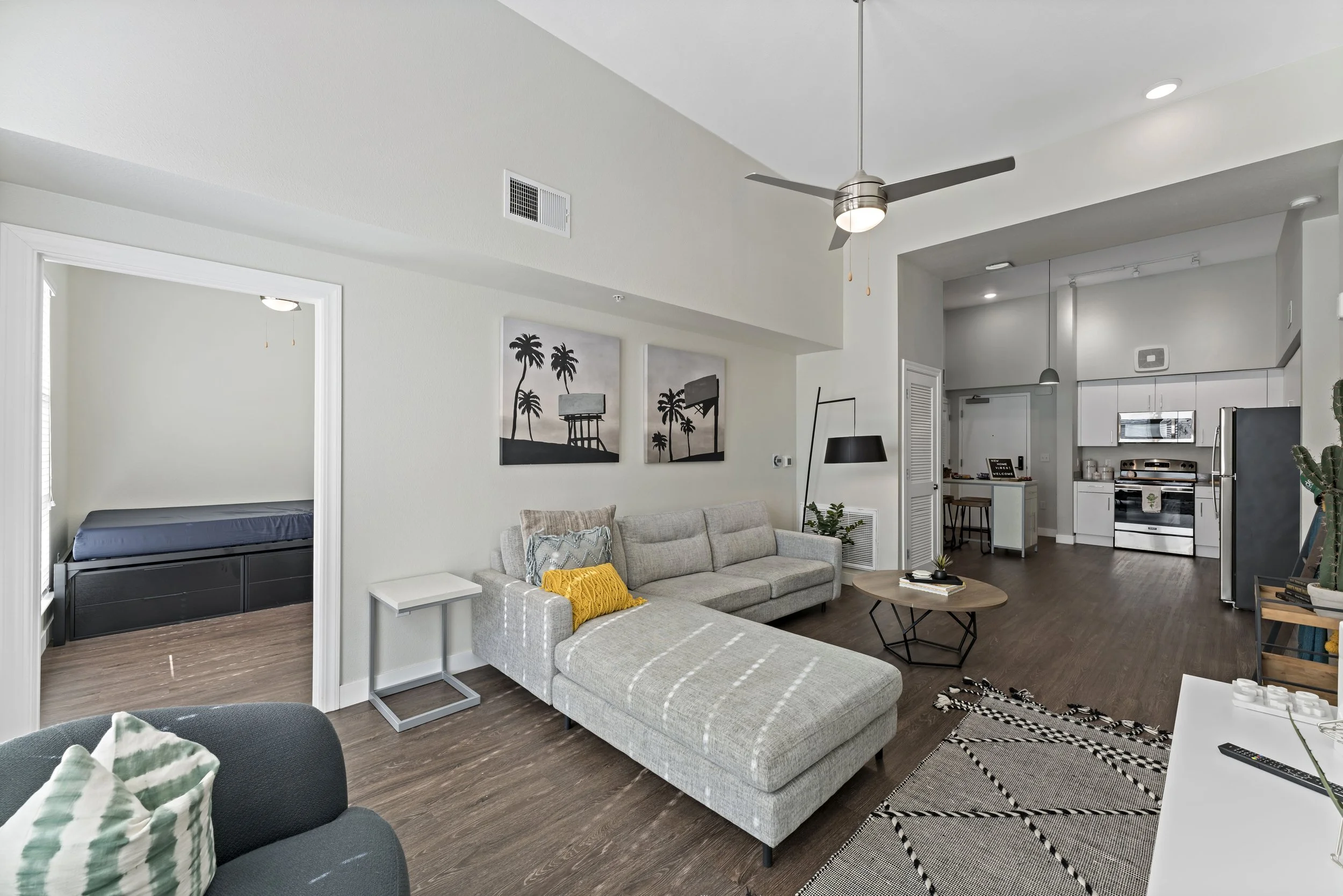 Living room with gray sectional sofa, black floor lamp, and wall art of palm trees, view of kitchen with white cabinets and stainless steel appliances, bedroom with bed, and decorative rug and plants.