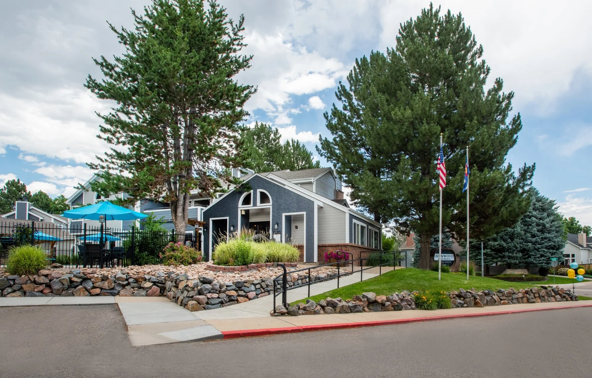 Residential neighborhood with a blue house, large trees, flagpoles with American and state flags, landscaped yard, sidewalk, and street.
