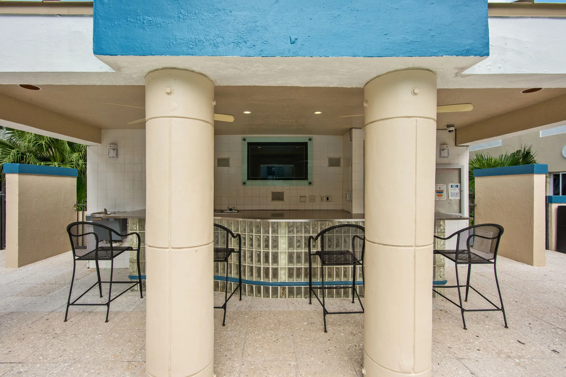 Outdoor bar area with four black metal chairs, a tiled countertop, a flat screen TV, and tropical plants in the background.