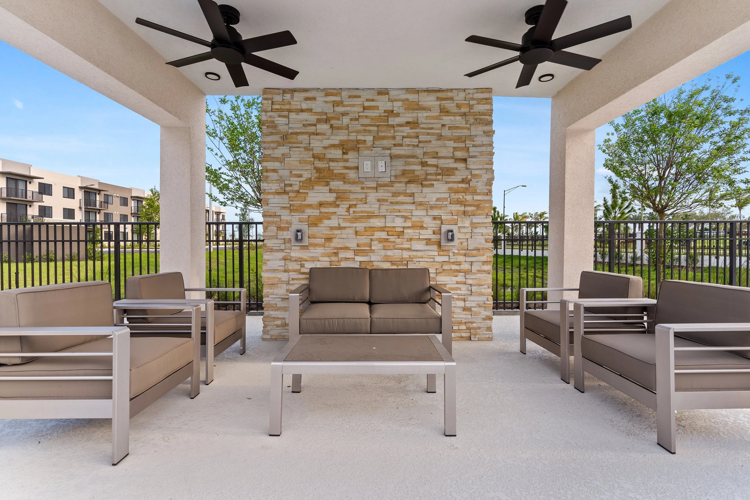 Outdoor covered seating area with brown cushioned chairs and a sofa, table, ceiling fans, and a stone accent wall, with a green park and blue sky in the background.