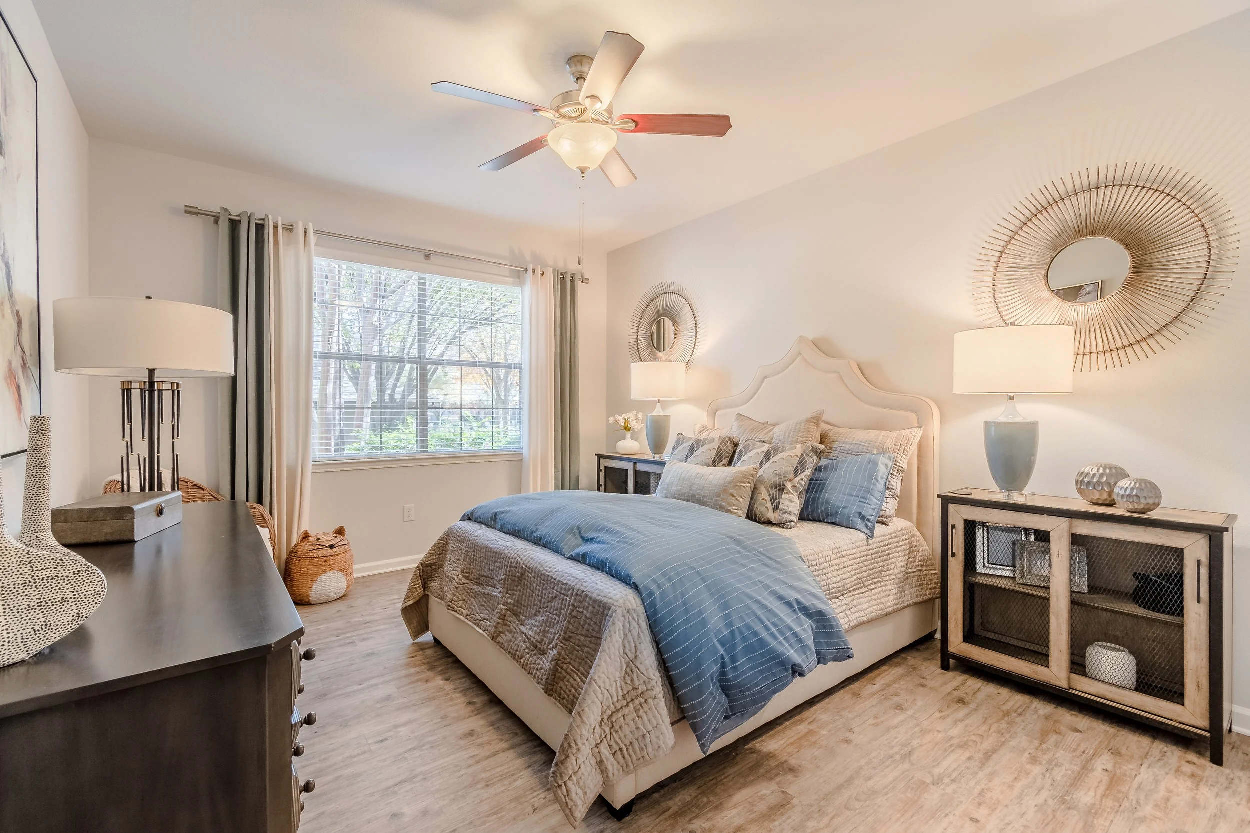 A cozy bedroom with a large window, a bed with blue and beige bedding, two nightstands with lamps, decorative wall mirrors, and a ceiling fan. The room has light-colored walls and wood flooring.