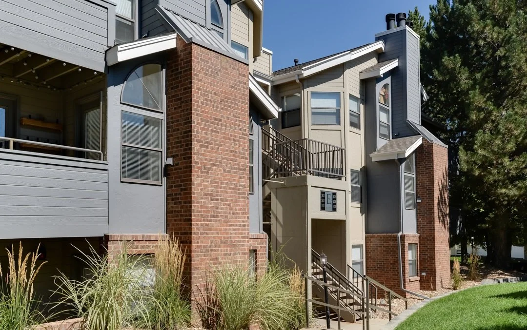 Exterior view of a modern apartment building with brick and gray siding, multiple windows, and stairways, surrounded by grass and trees.