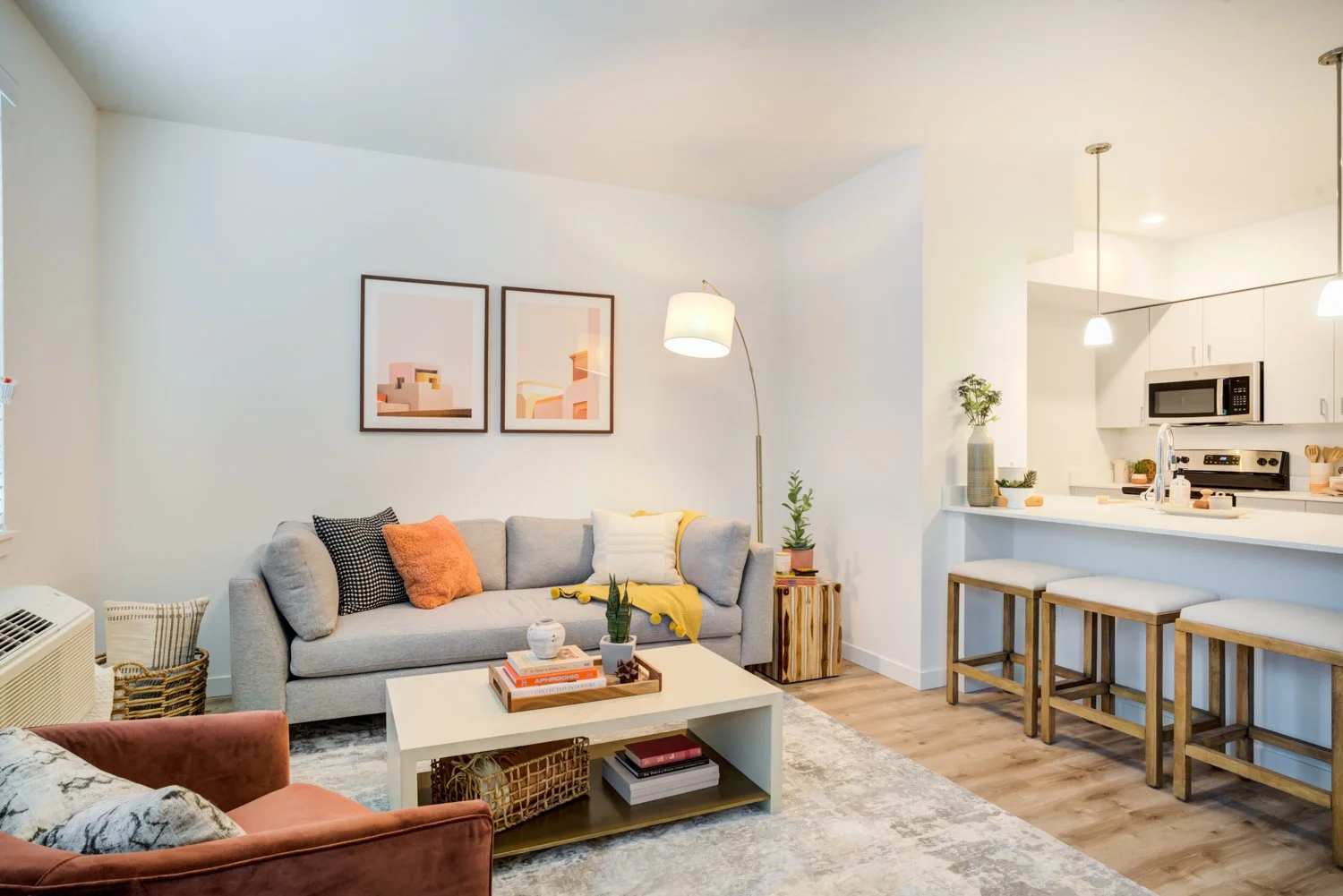 A cozy living room with a light gray sofa decorated with orange, black, and white pillows, a white coffee table with books and plants, and a patterned area rug. There are two framed abstract artworks on the wall, a tall floor lamp, and a side table w