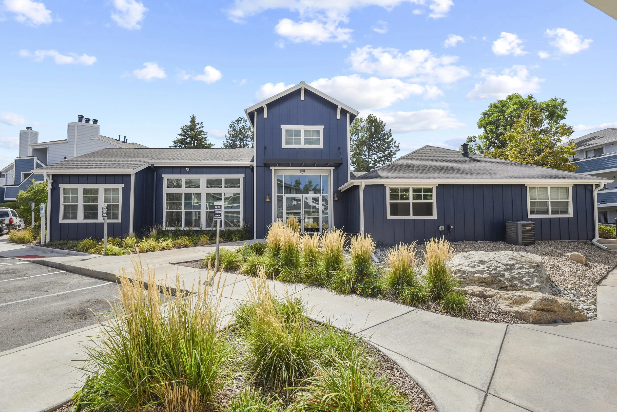A two-story blue building with white trim, large windows, and a small entrance, surrounded by a well-maintained garden with ornamental grasses, rocks, and a sidewalk under a partly cloudy sky.