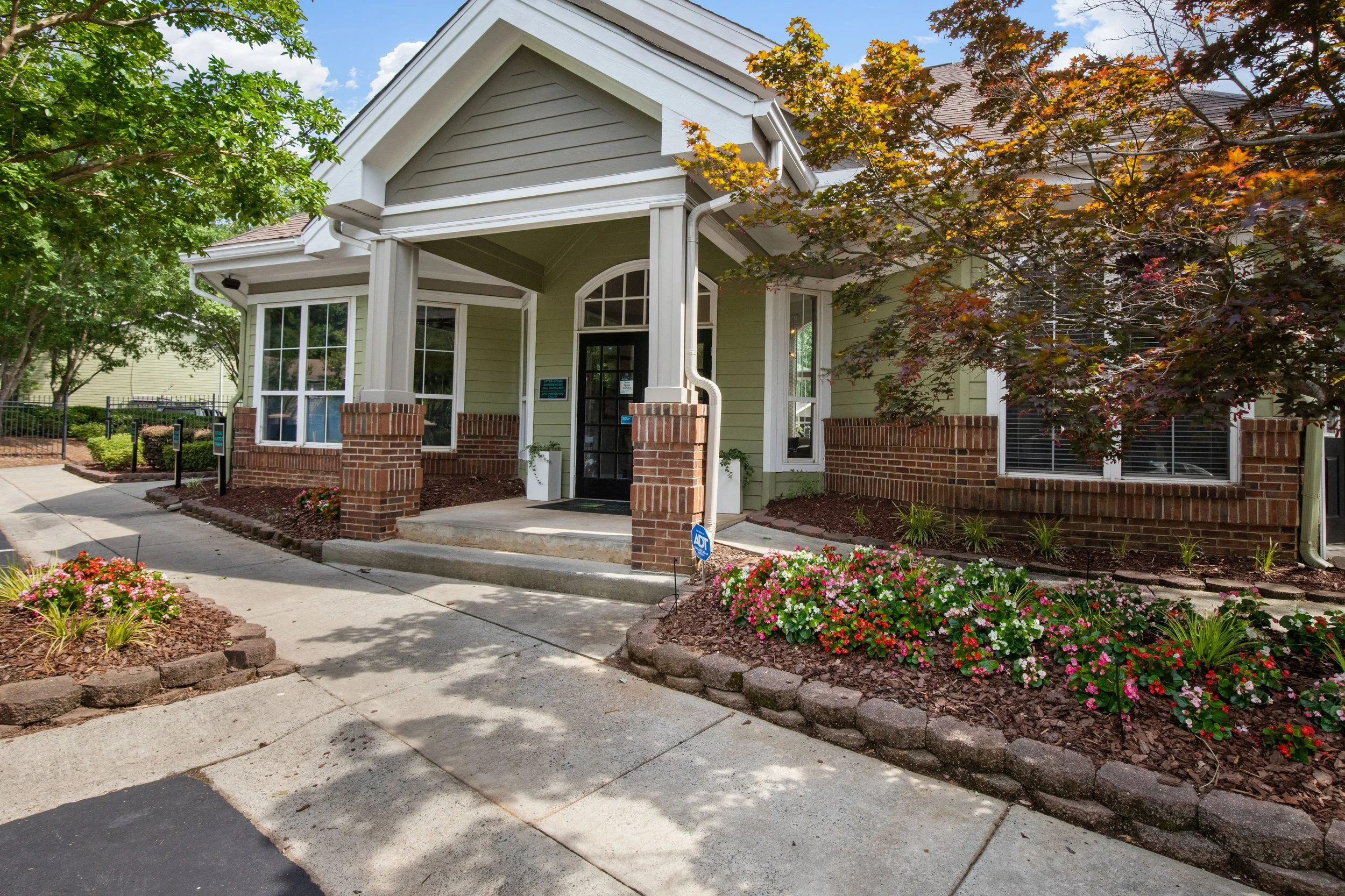 A modern residential house with a light green exterior, brick accents, large windows, and a covered front porch with brick columns. There are colorful flower beds and trees along the sidewalk under a partly cloudy sky.