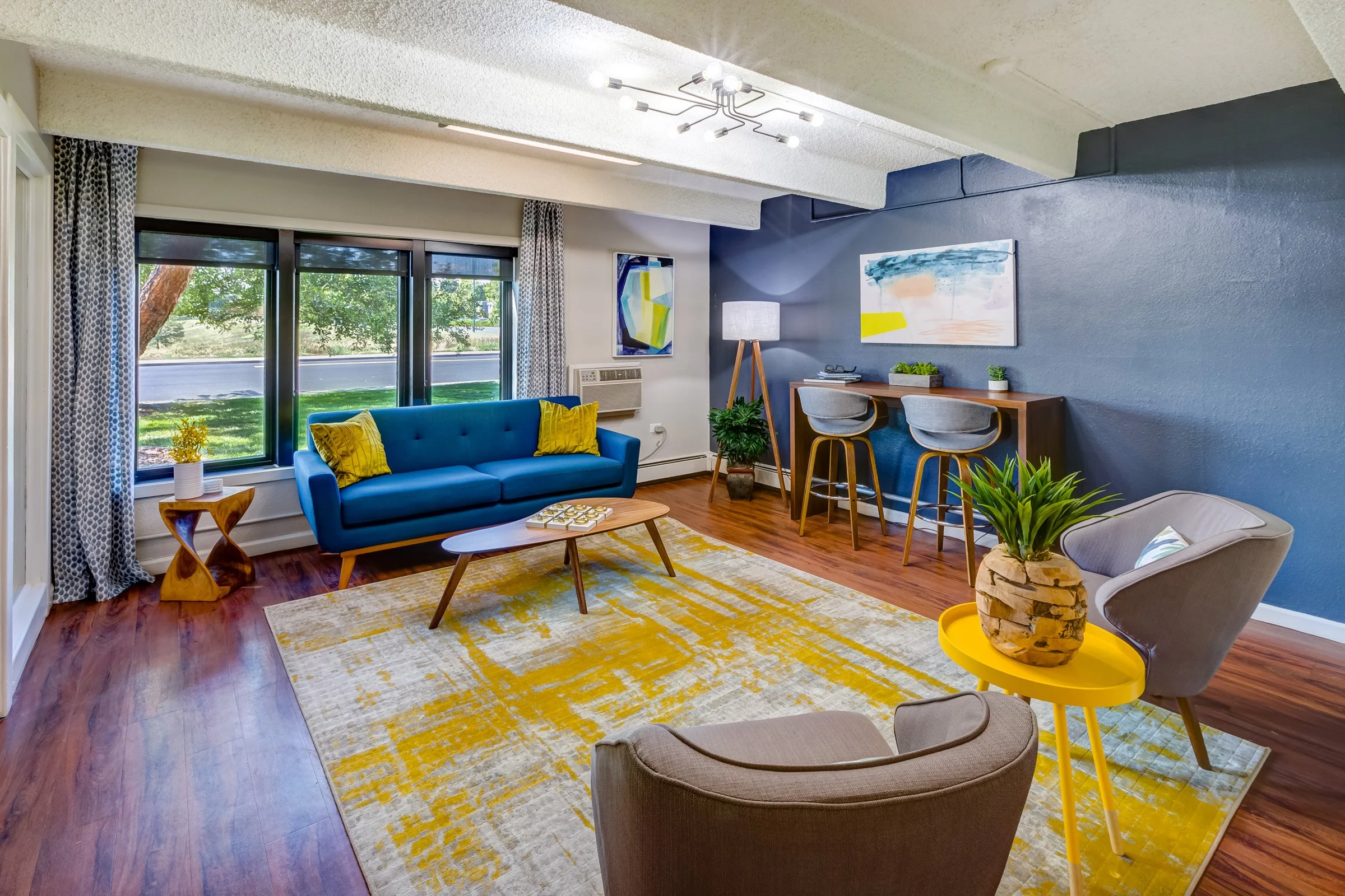 Living room with a blue couch, beige armchair, and yellow side table, large window with curtains, wooden floor, and decorative plants.