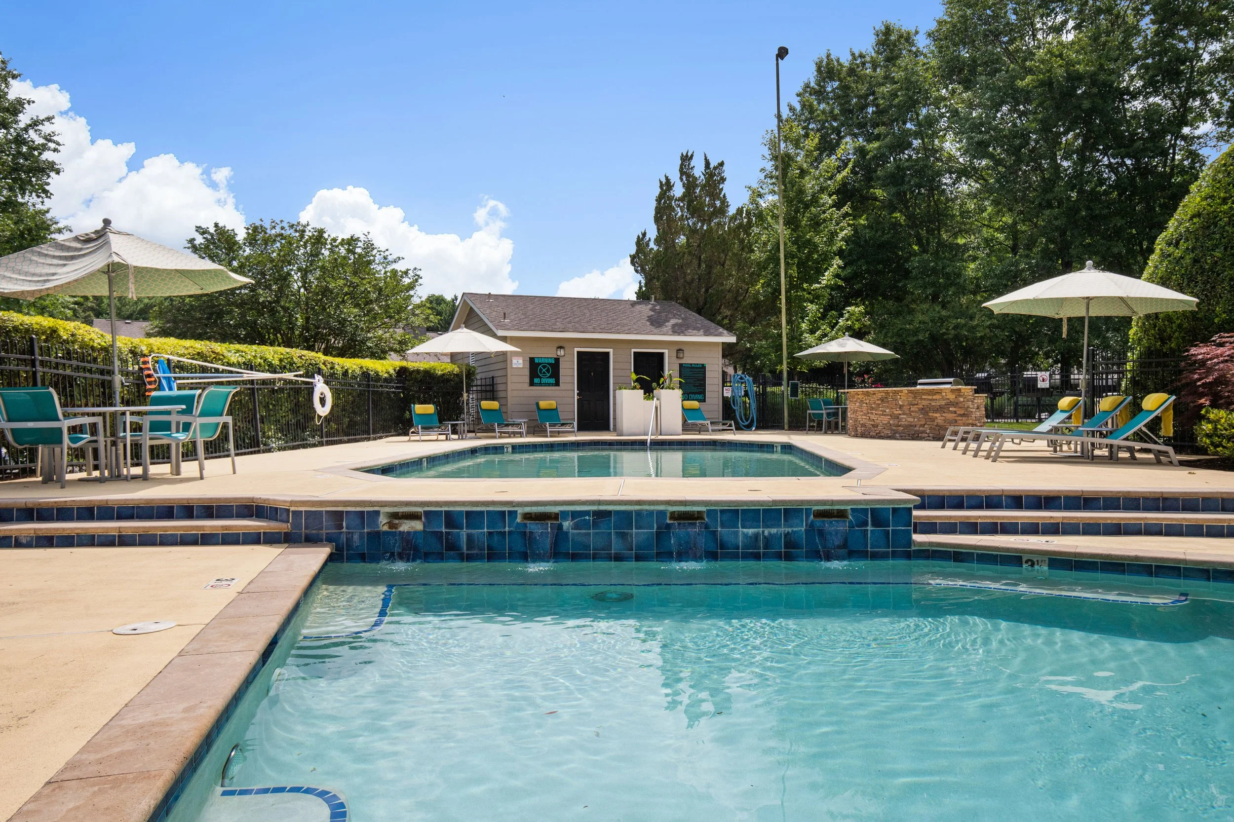 Outdoor swimming pool with lounge chairs, umbrellas, a small pool house, and lush green trees under a partly cloudy sky.