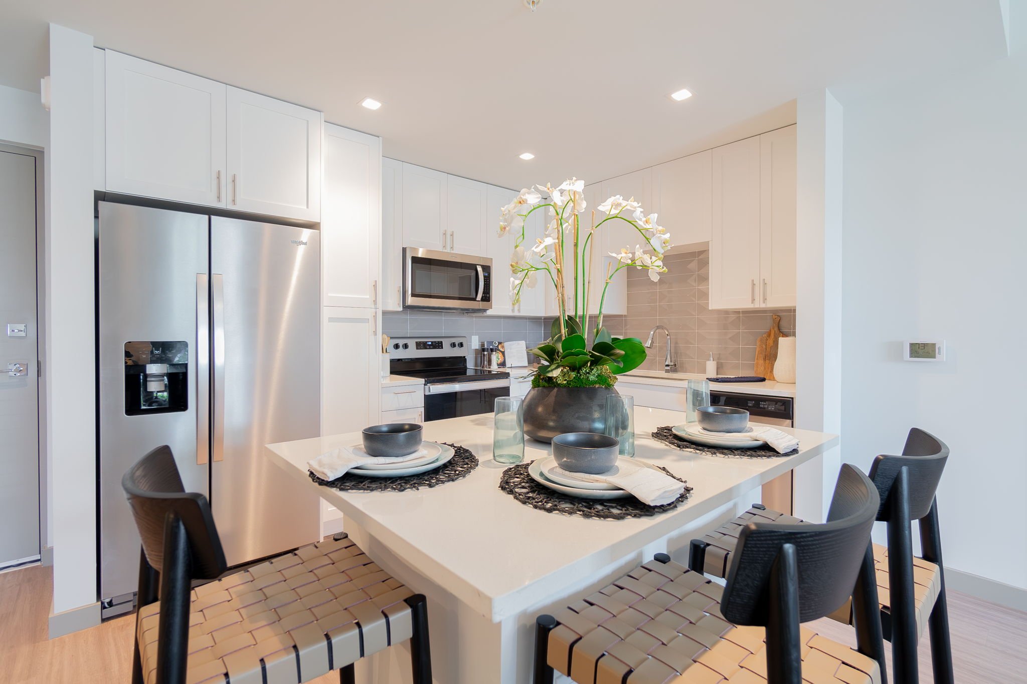 Modern kitchen with white cabinets, stainless steel refrigerator, microwave, and oven. Center island with a white countertop, decorated with a potted orchid and table settings for four, including black bowls, white plates, and napkins. Four black cha