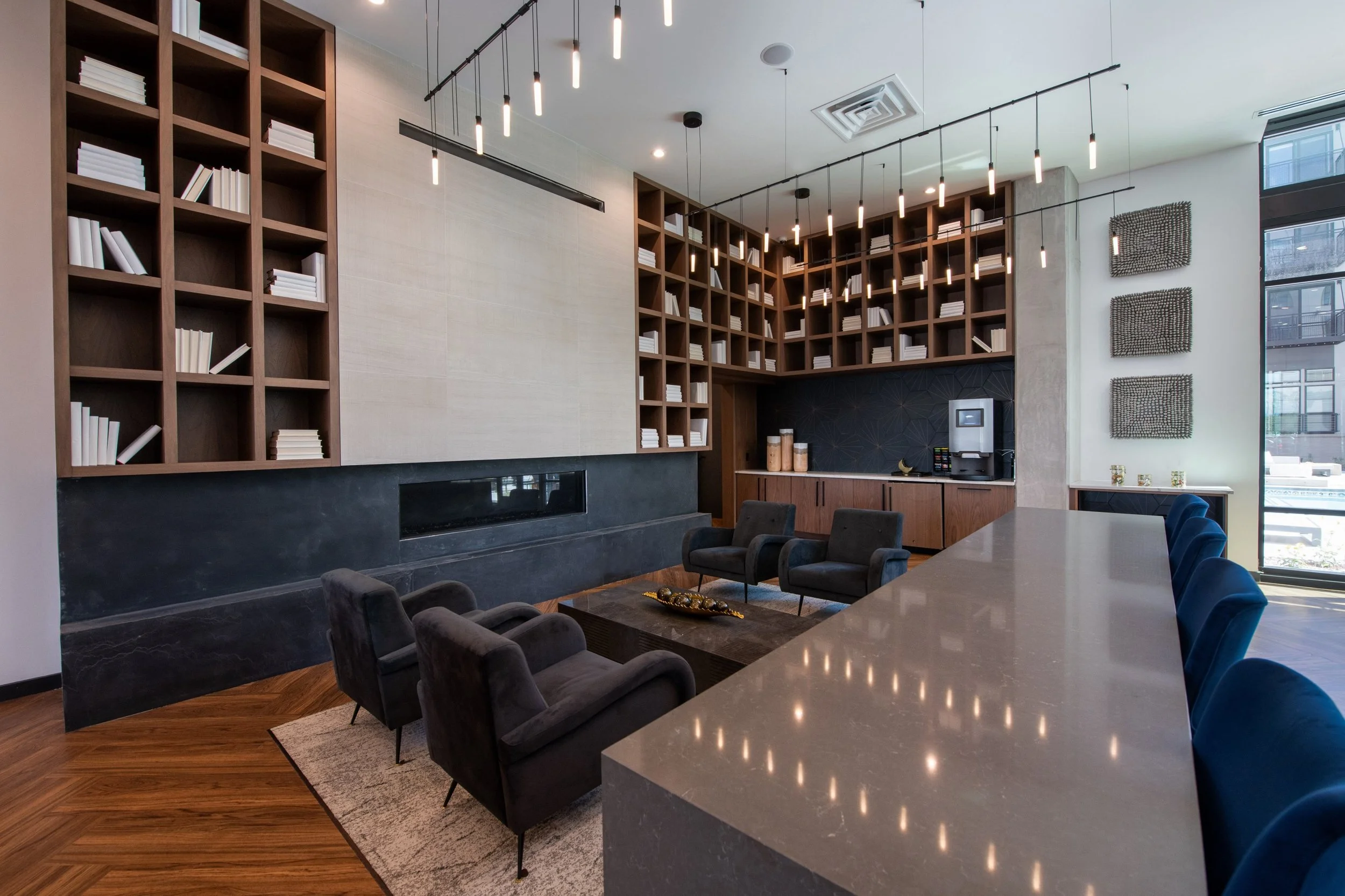 Modern lounge area with a large beige countertop, black chairs, dark wood and beige bookshelves, pendant lighting, and a coffee station.