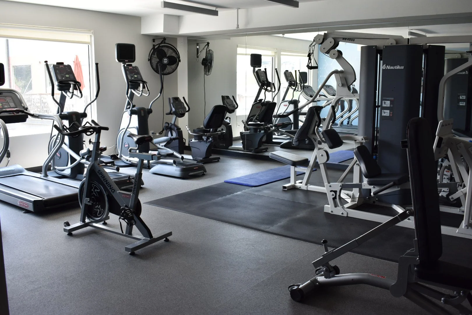 Empty gym room with various cardio and weight machines, large mirror on the wall, and windows with natural light.