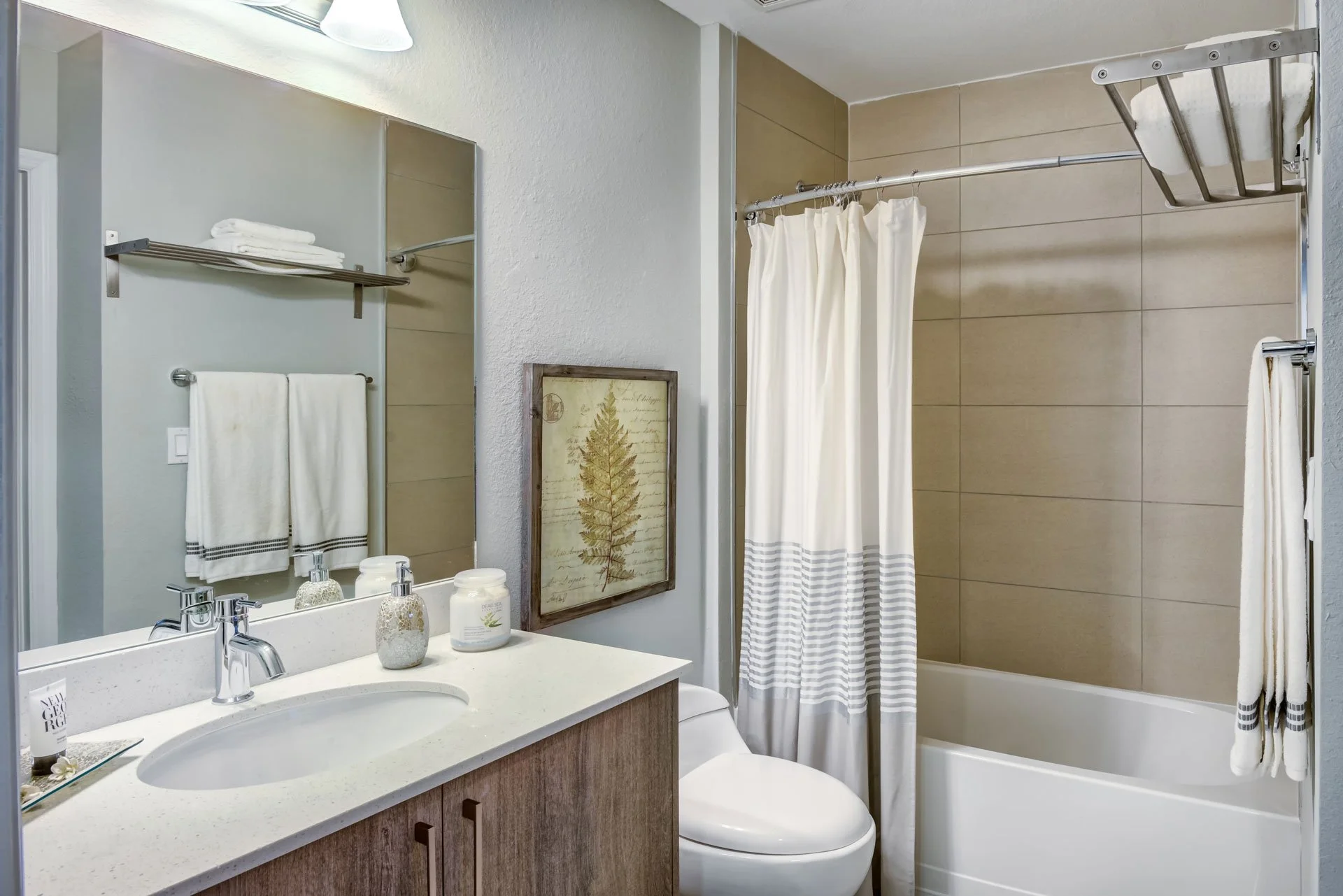 Bathroom with vanity, mirror, and sink on the left, towels hanging on a rack above, and a framed botanical art piece on the wall. To the right, there is a toilet and a bathtub with a shower curtain, tiled walls, and a metal shower rack.