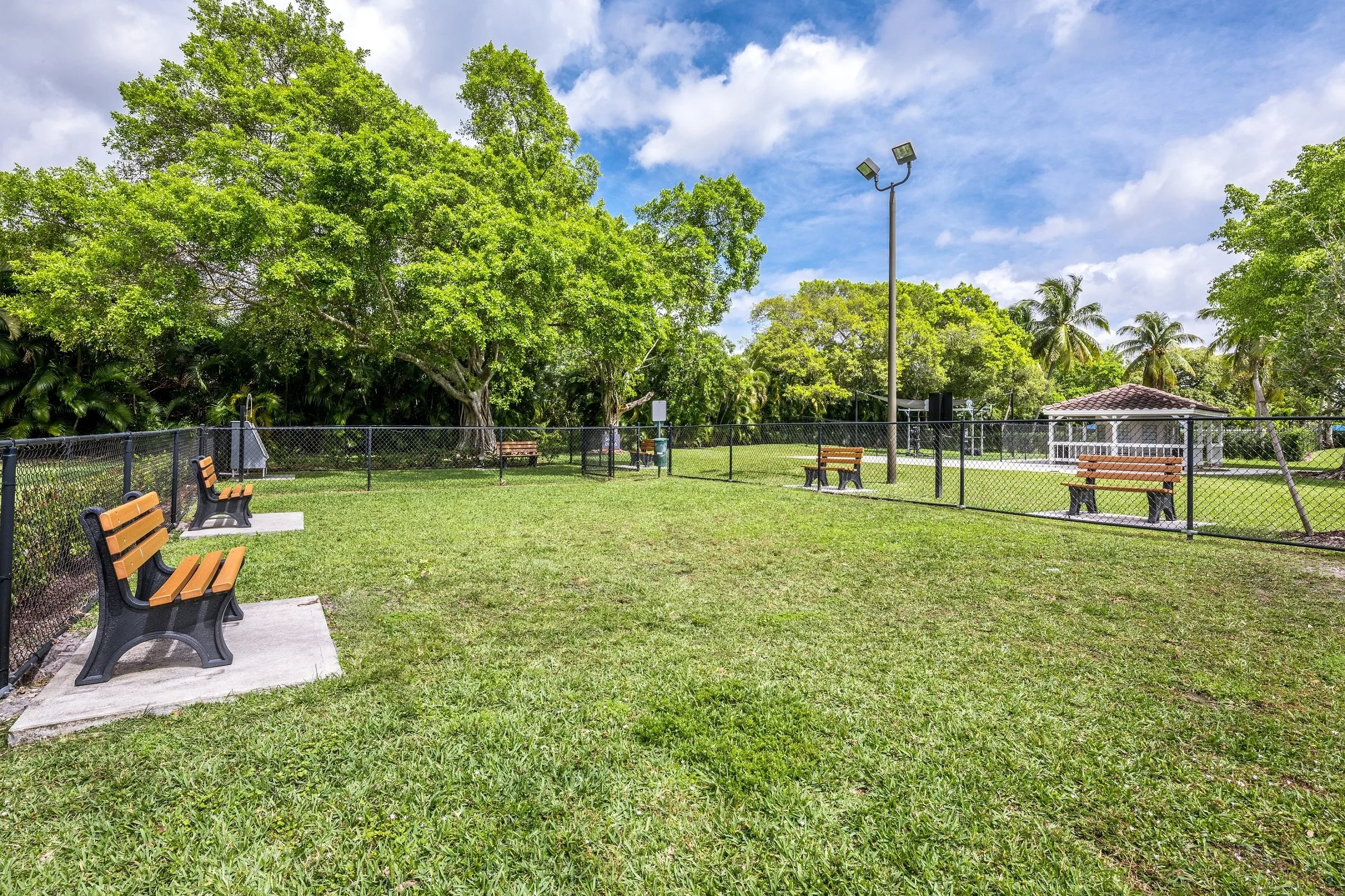 A fenced grassy park area with benches, trees, a small pavilion, and a tall light pole under a partly cloudy sky.
