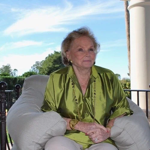 An elderly woman sitting outdoors on a cushioned chair, wearing a shiny green blouse and gold earrings, with trees and a blue sky in the background.