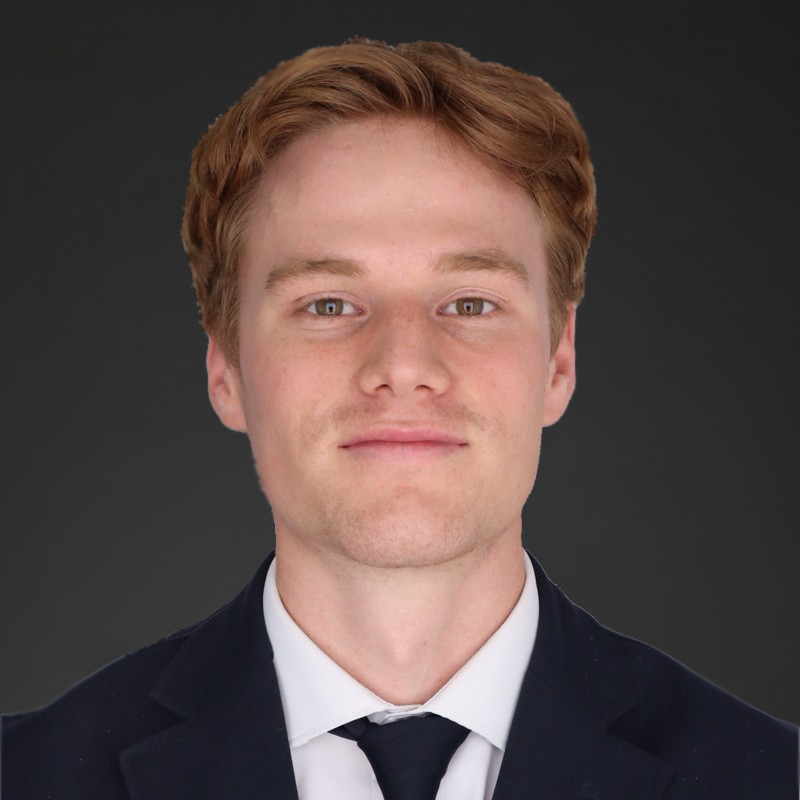 A young man with red hair, wearing a dark suit, white shirt, and black tie, against a gray background.