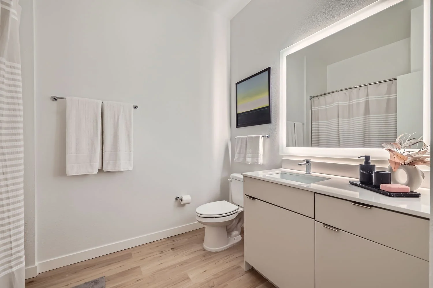 Modern bathroom with white walls, a large backlit mirror, a white vanity with a modern faucet, and a toilet. There are white towels on a metal rod, a black tray with toiletries, and a decorative vase with pink leaves. A framed landscape painting hang