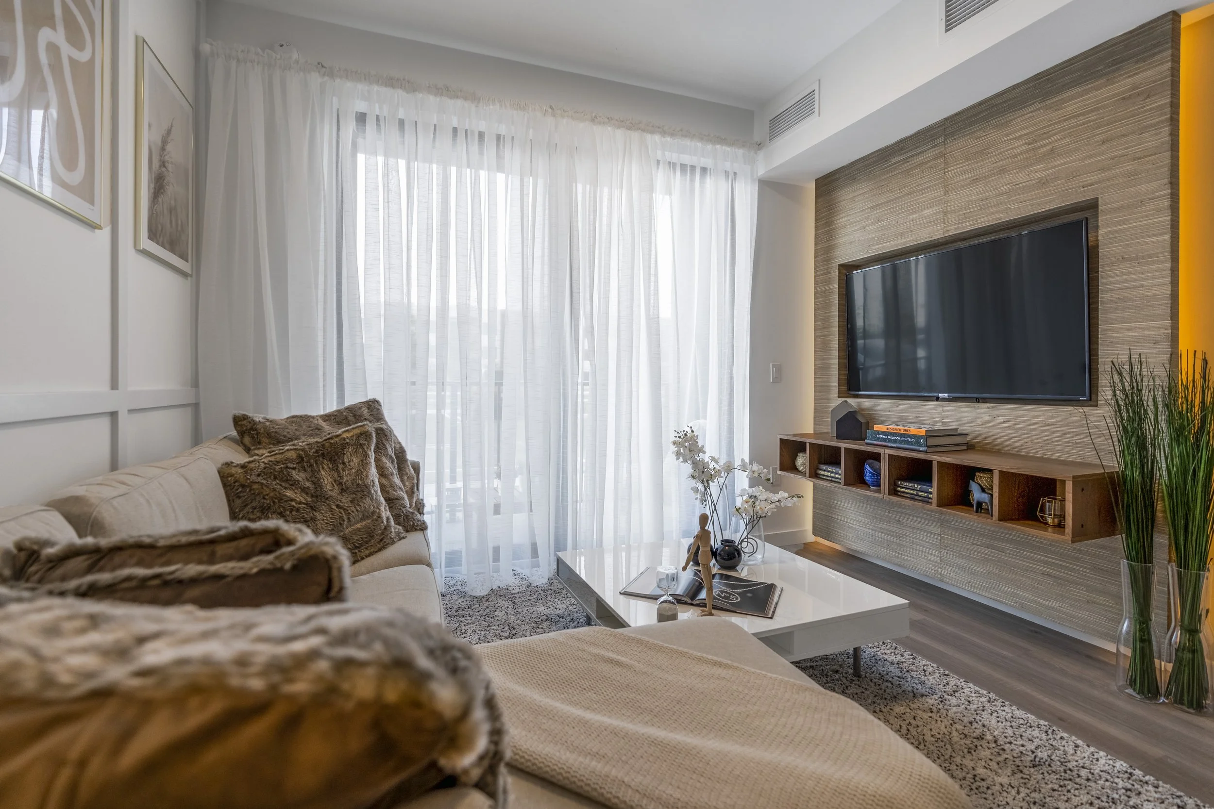 Modern living room with beige sofa, fur pillows, sheer white curtains, a flat-screen TV on a wood-paneled wall, decorated with books and plants on a floating shelf.