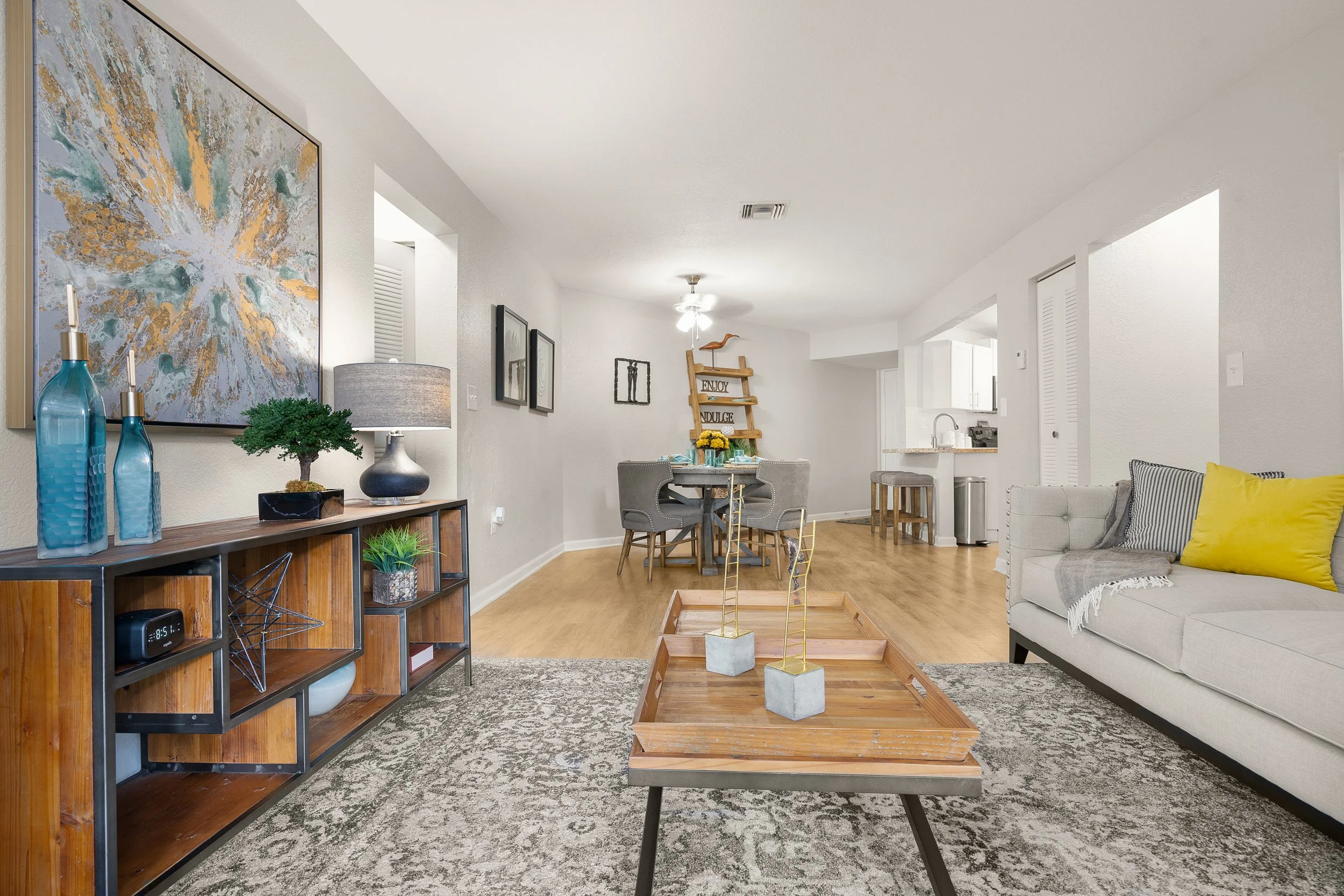 Living room with beige sofa, yellow and striped pillows, wooden coffee table, art on walls, and a dining area with gray chairs and a decorative ladder with signs.