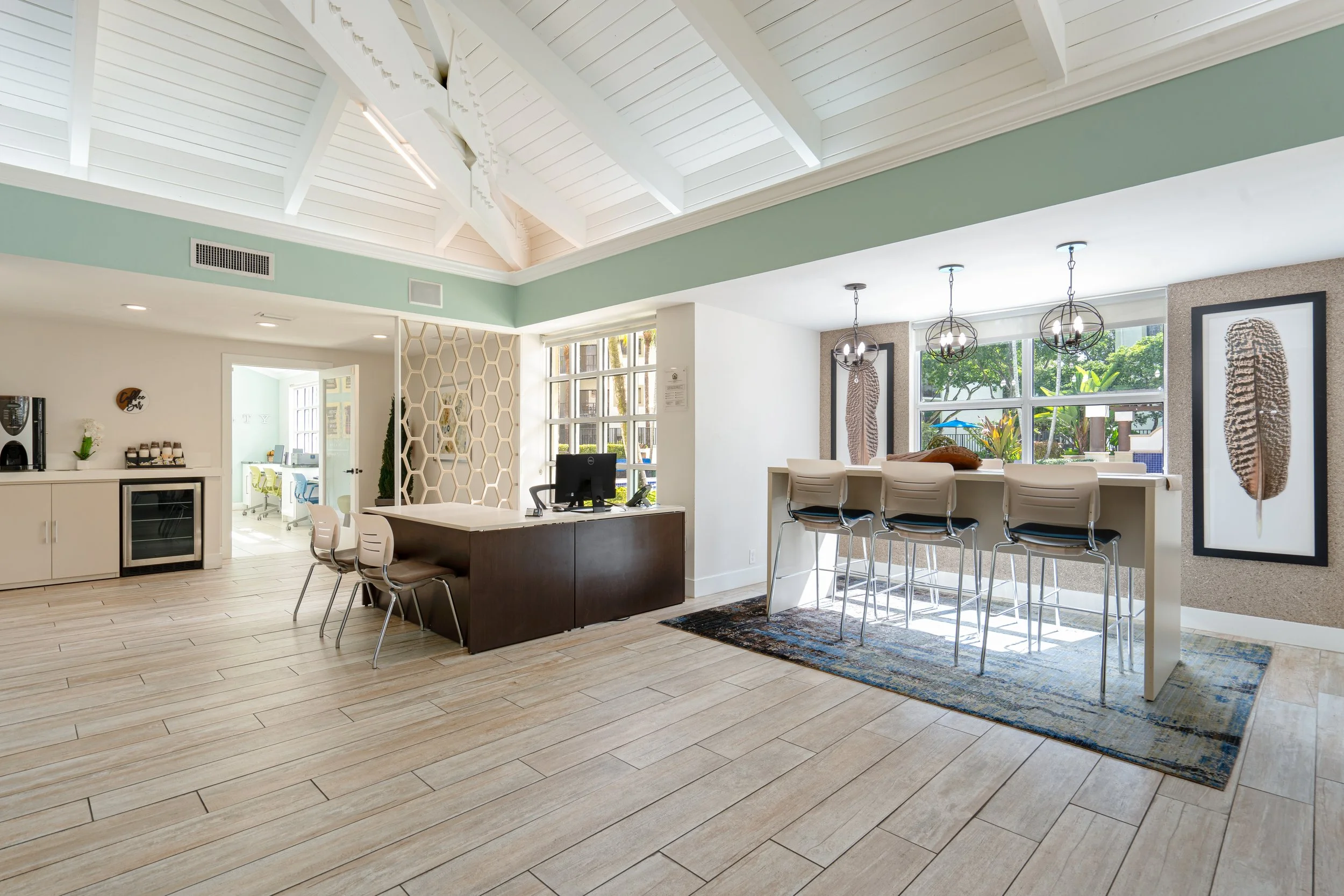 Modern hotel lounge with high ceiling, bar-height table with four chairs, large windows with greenery outside, and framed art of feathers on the wall.