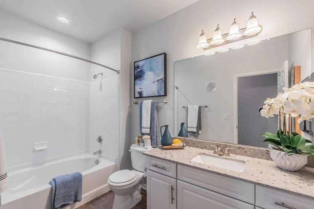 A modern bathroom with white tiles and a bathtub on the left, a toilet in the middle, and a white vanity with a granite countertop, a mirror, and decor on the right. There are towels hanging and flowers on the counter.