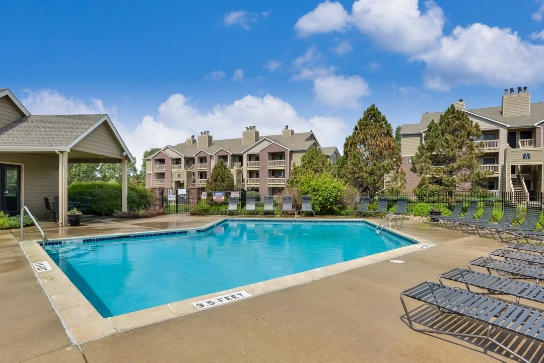 Community swimming pool with lounge chairs, surrounded by apartment buildings and greenery on a sunny day.