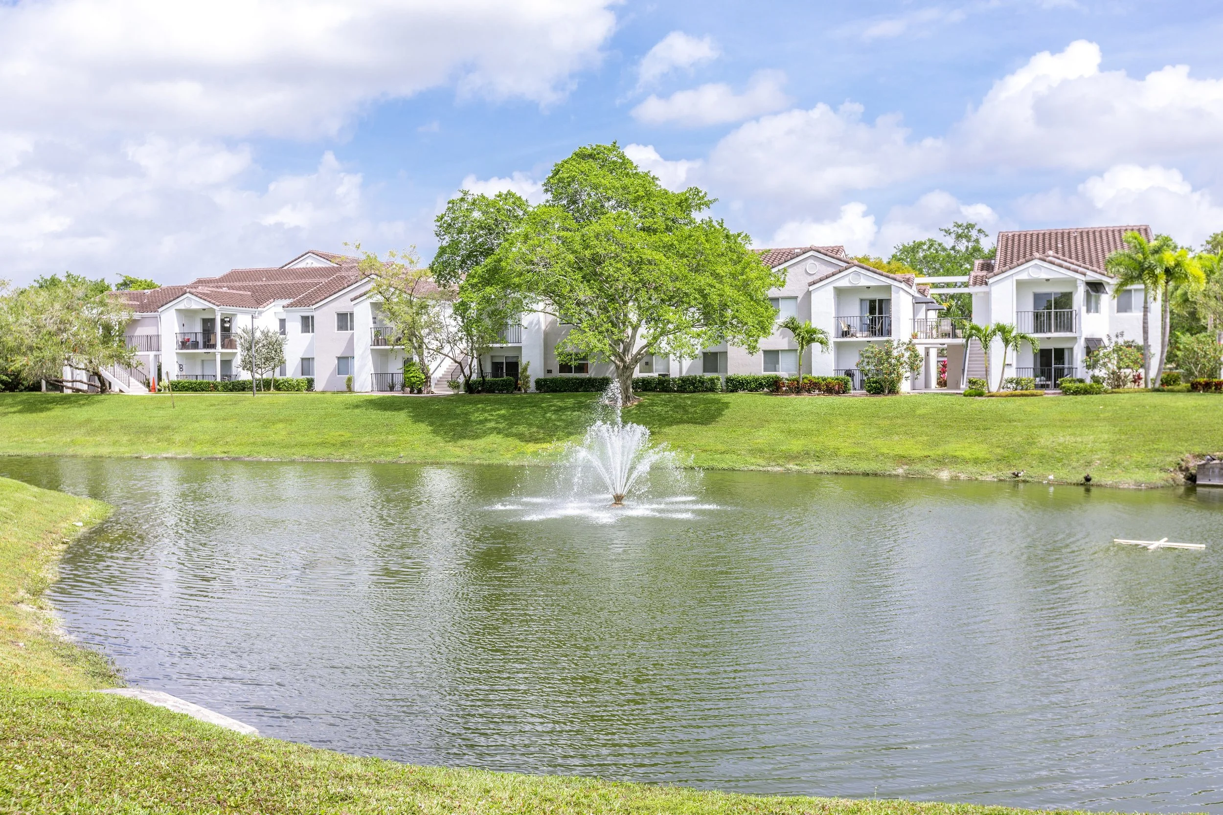 Apartment complex with white buildings, balconies, and red-tiled roofs, situated behind a pond with a fountain, green grass, trees, and a partly cloudy sky.