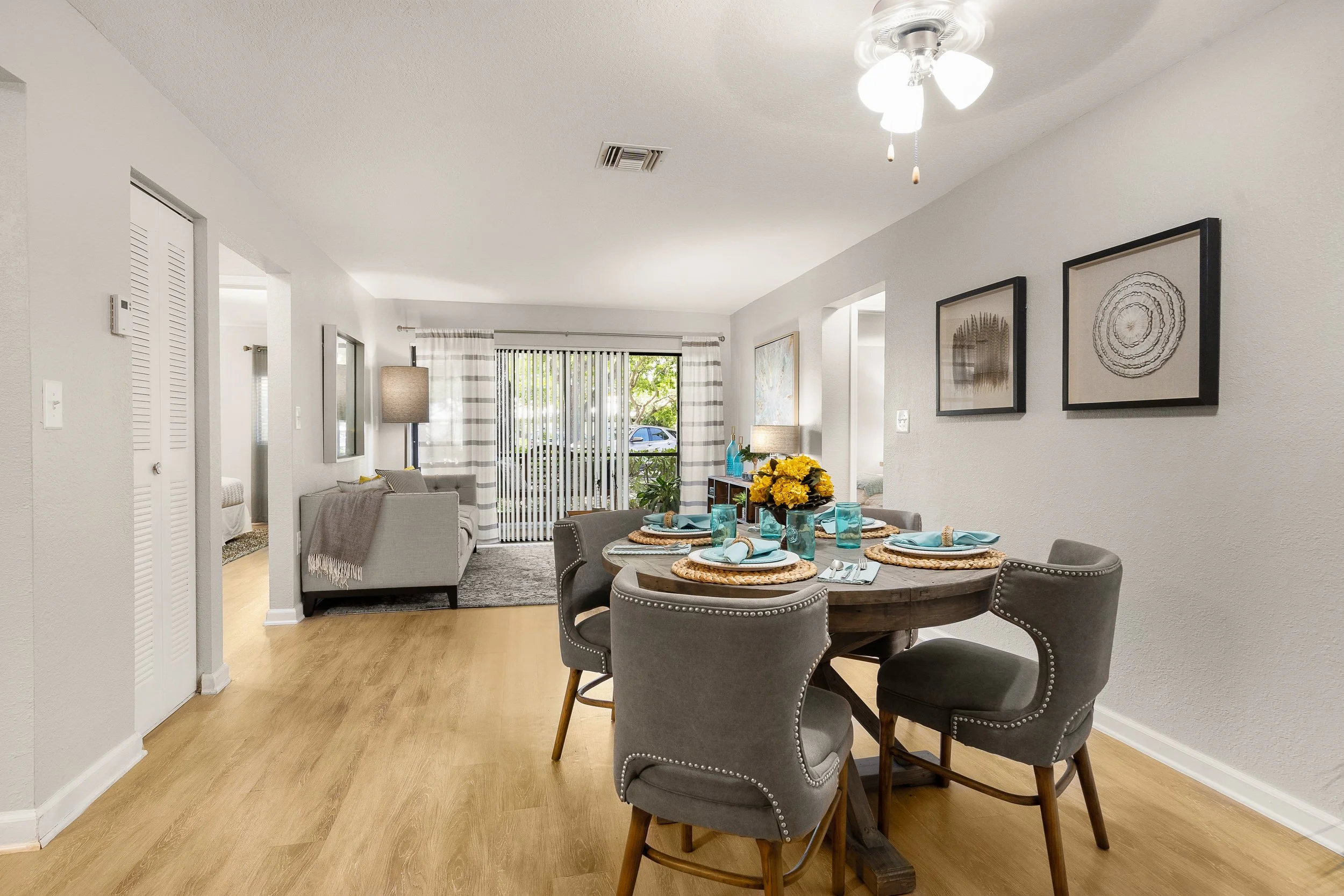 Living and dining area in a modern home with beige walls, wood flooring, a round dining table with four gray chairs, a gray sofa, artwork on the walls, and a sliding glass door with striped curtains leading outside.