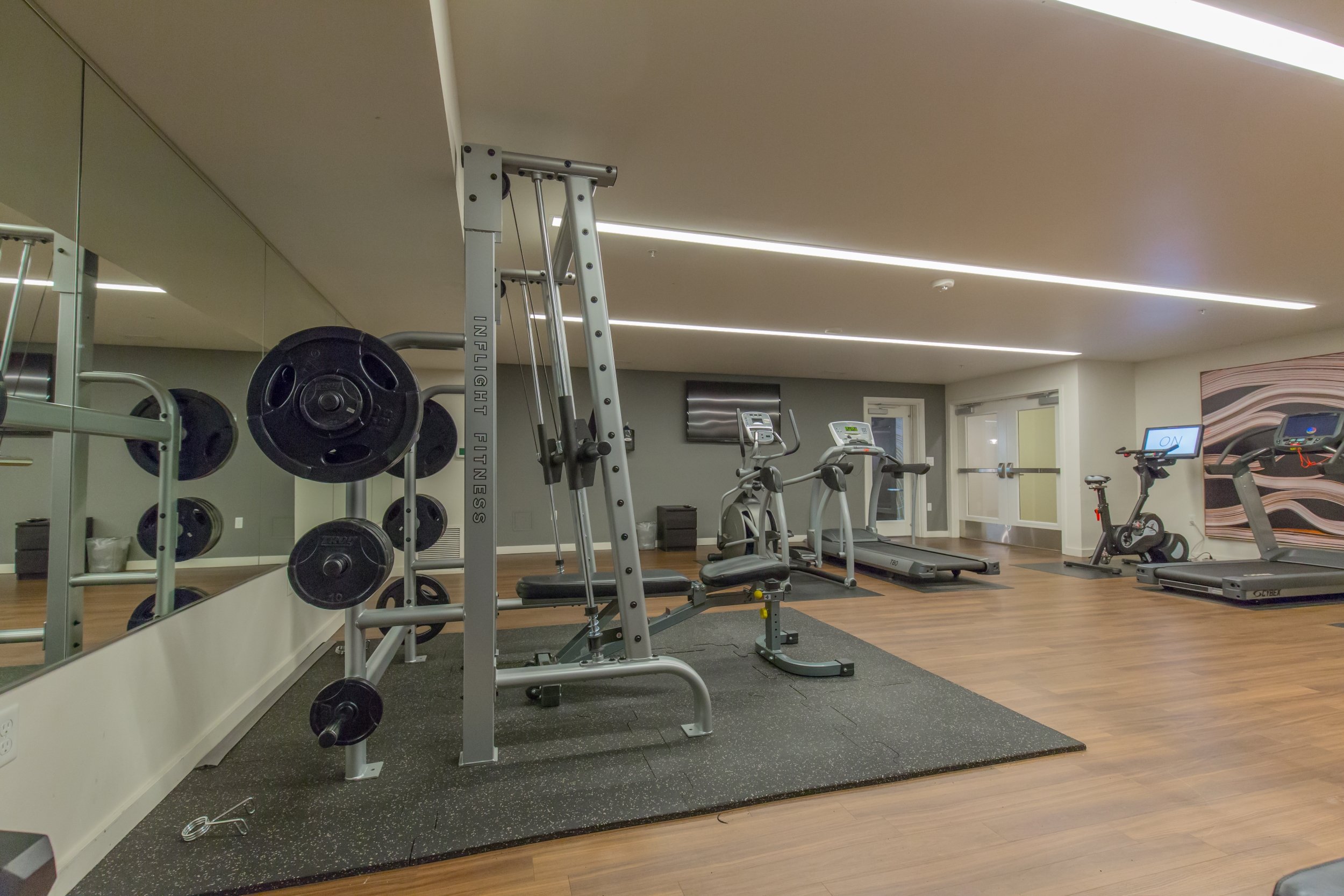 Empty gym with various exercise equipment, including weights, treadmill, stationary bike, and elliptical, on wood flooring with mirrored wall.