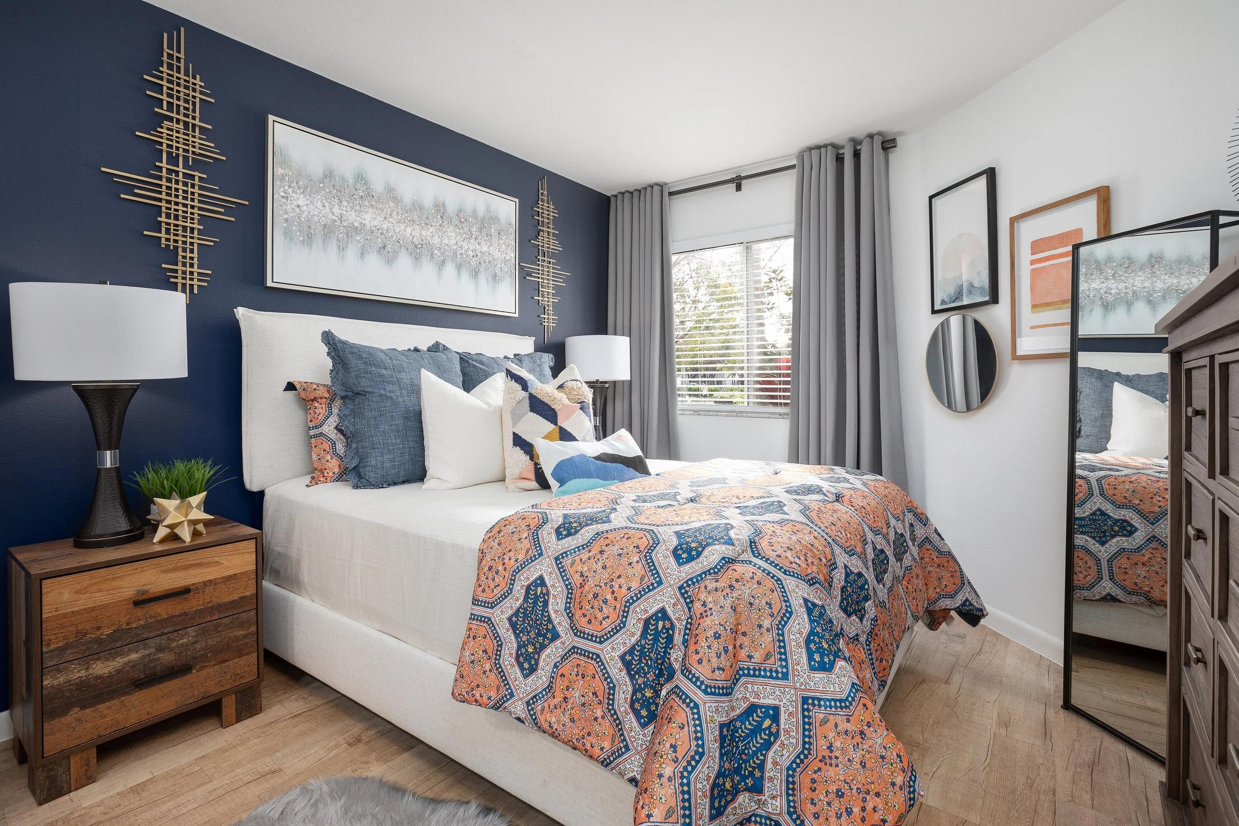 Modern bedroom with a white bed, colorful patterned bedspread, and decorative pillows. Blue accent wall with wall art, wooden nightstand with a lamp, and window with gray curtains. Full-length mirror and framed artwork on white wall.