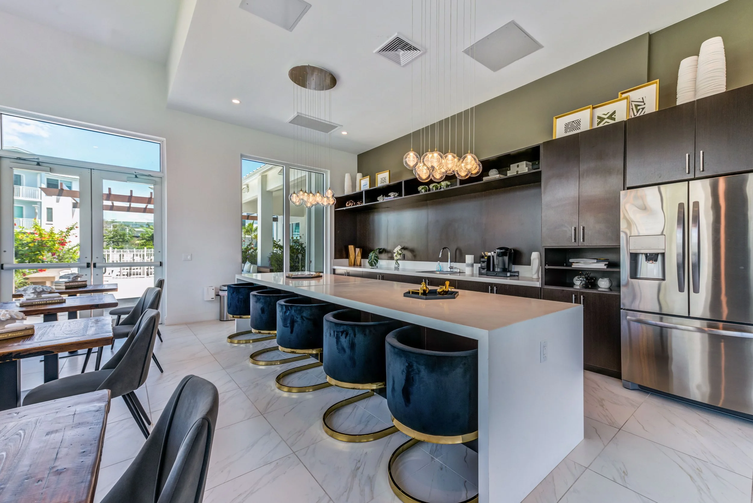 Modern kitchen with white island, five blue velvet barstools with gold bases, dark wood cabinets, stainless steel refrigerator, and dining area with wooden tables and gray chairs, large windows and glass doors