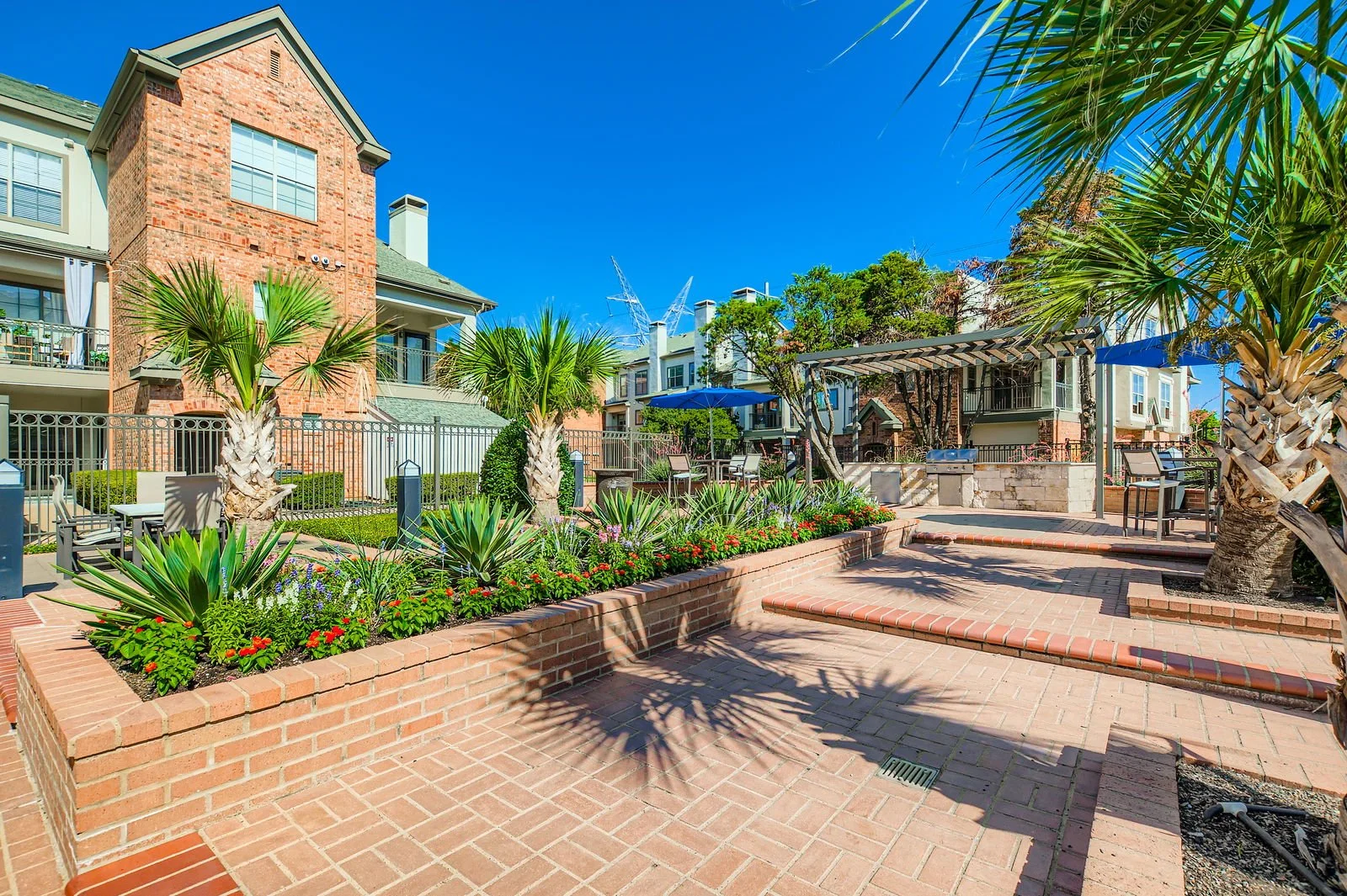 Outdoor courtyard with brick paving, tropical palm trees, colorful flower beds, tables with umbrellas, a built-in barbecue area, and residential buildings in the background under a clear blue sky.