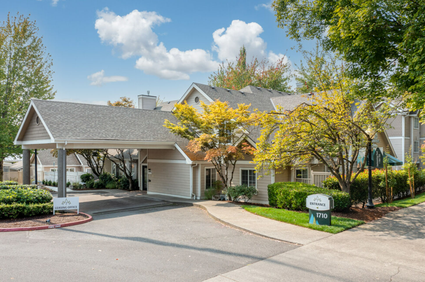Image of a residential building with a driveway, trees, and signs indicating leasing office and entrance, surrounded by landscaped bushes and a sidewalk under a partly cloudy sky.