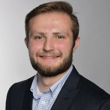 Professional headshot of a young man with brown hair and a beard, wearing a dark suit jacket and light blue shirt, smiling against a plain gray background.