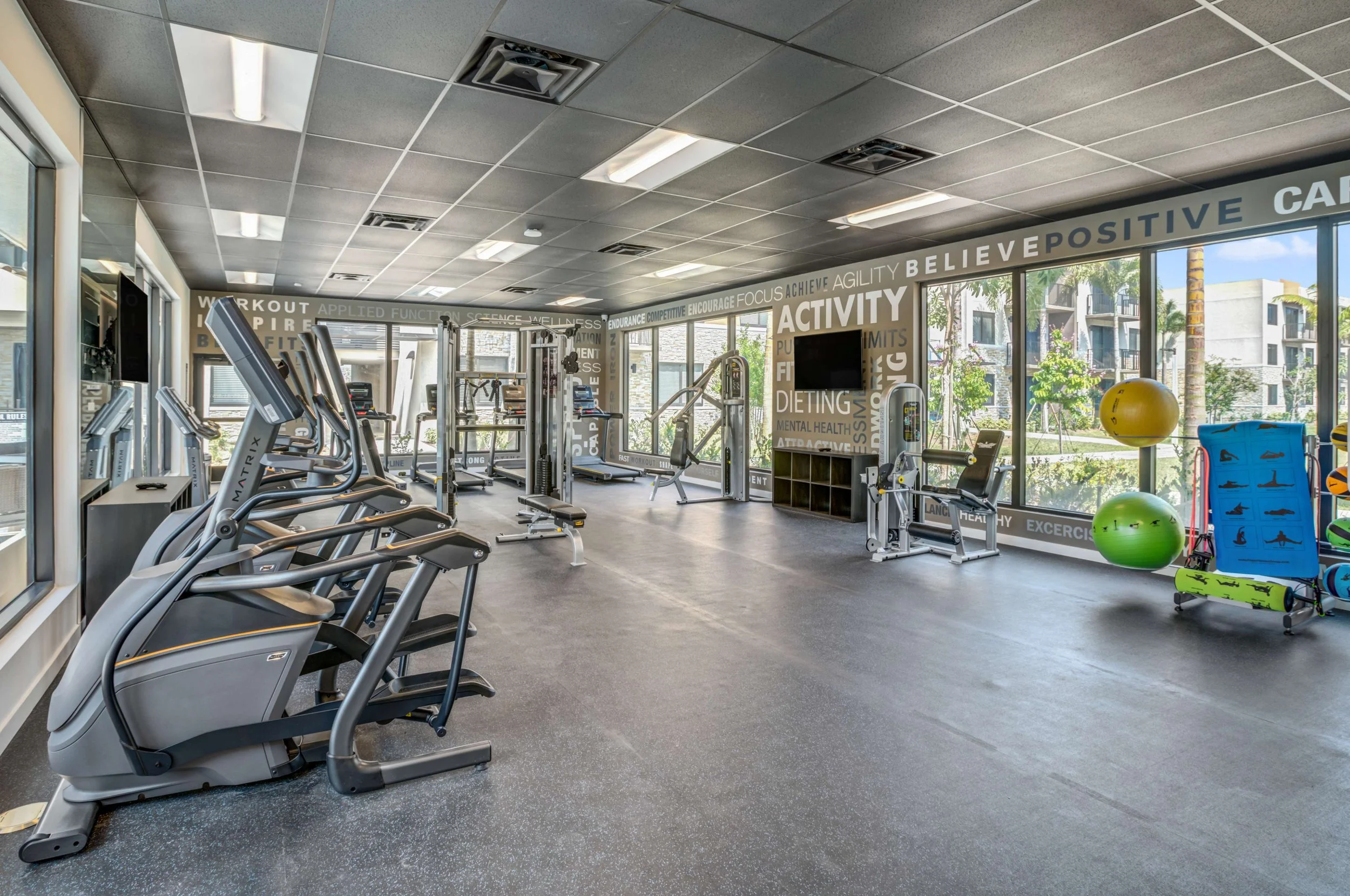 Empty gym with workout equipment, exercise machines, stability balls, and bright large windows with a city view.