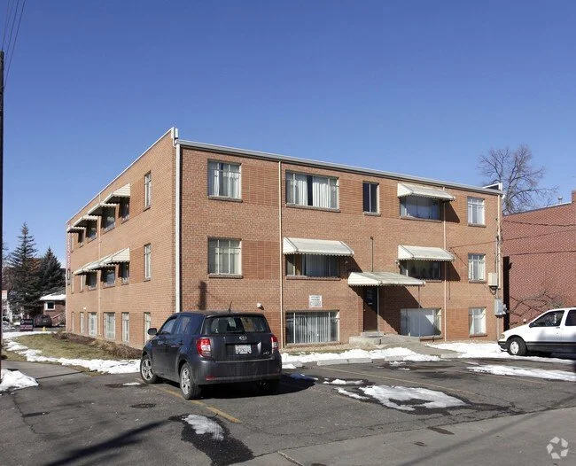 A three-story brick apartment building with three awnings over the entrance and windows, parked cars in front, patches of snow on the ground, and a clear blue sky.